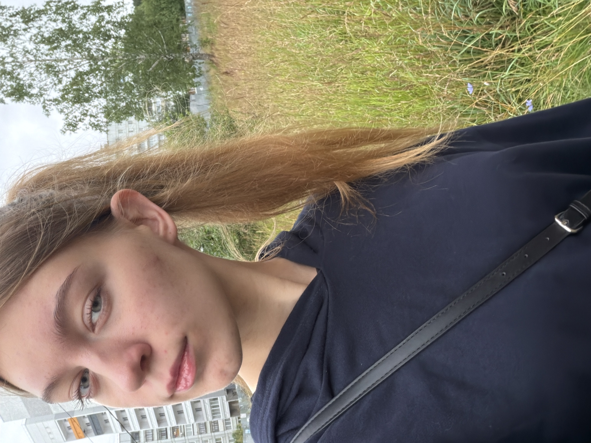 A young woman with blonde hair pulled into a ponytail poses for a selfie outdoors. She is wearing a dark blue hoodie and a black crossbody bag. Behind her, a tall tree stands in a field of tall grass. In the background, a multi-story apartment building is visible, suggesting an urban or suburban setting. The sky is overcast. The specific location in Chaikovsky, Russia, is not discernible from the image alone.