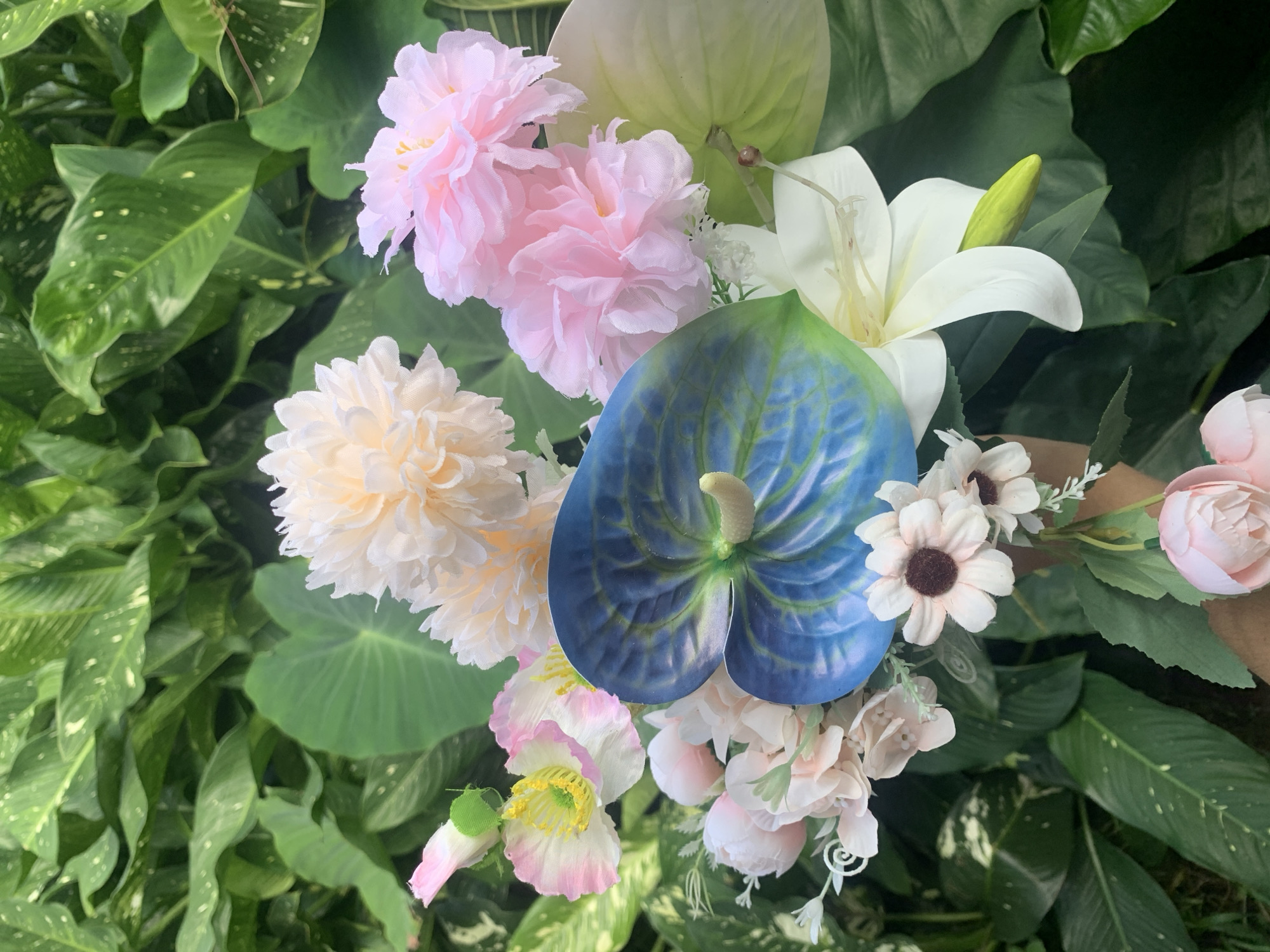A close-up, overhead shot captures a bouquet of artificial flowers arranged against a backdrop of lush green foliage. The bouquet features a prominent blue and green anthurium, a white lily, various shades of pink and cream-colored ruffled flowers, and smaller white flowers with dark centers. The foliage appears to be a mix of broad, waxy leaves, some with white speckles. The lighting suggests an outdoor setting, possibly a garden or park. No individuals or specific landmarks are visible, so a precise location within Kuala Pembuang, Indonesia, cannot be determined from the image.