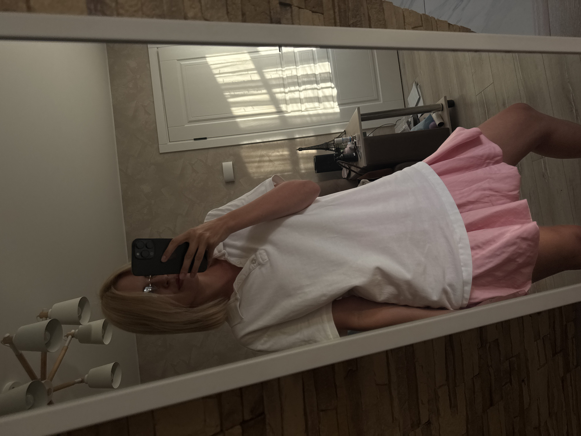 A person is taking a mirror selfie in what appears to be a room. They are wearing a white t-shirt and a pink skirt. The person's hand holding a black smartphone is visible. In the reflection, a wooden floor and a wall with a textured pattern are visible. A closed white door with blinds casting shadows is also in the reflection. A lamp with multiple light fixtures is to the left of the person, and a shelf with items, including a small Eiffel Tower replica, is further back. The lighting suggests it might be daytime. The overall impression is a casual, personal moment captured in a domestic setting.