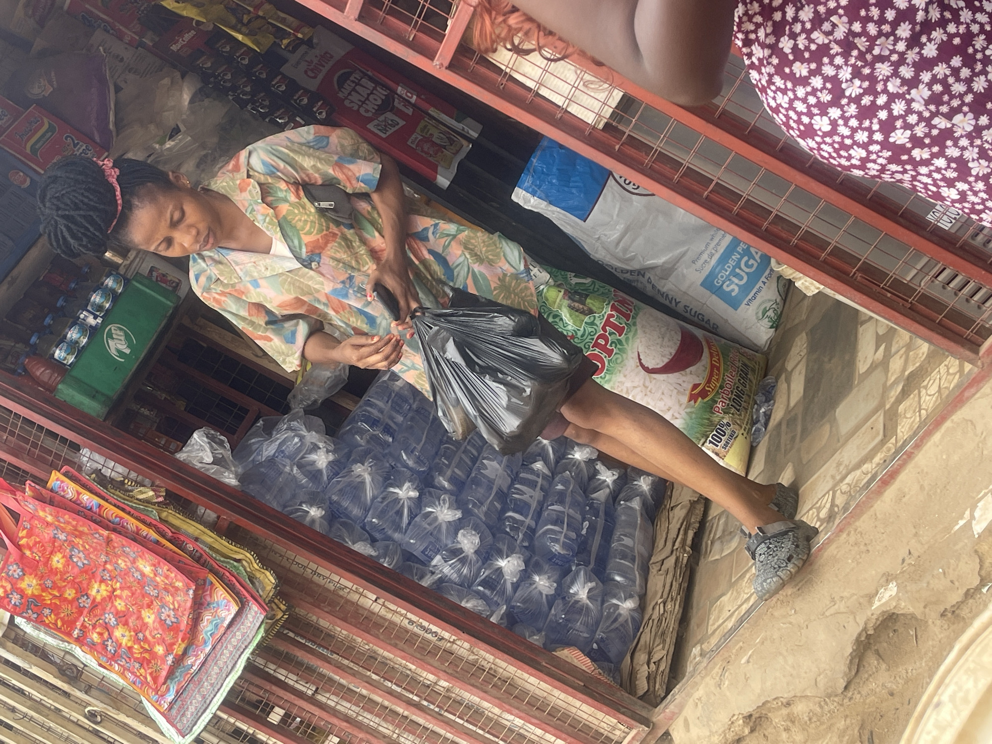 A woman with dark hair and a pink hairband stands inside a small shop in Abuja, Nigeria. She is wearing a patterned shirt with orange and green hues. She holds a black plastic bag. She is facing bags of sachet water and a bag of rice. Behind her, the shelves are stocked with food items. In the foreground are piles of reusable tote bags. Above, the upper level of the shop has bags of refined sugar and a woman with a patterned dress.