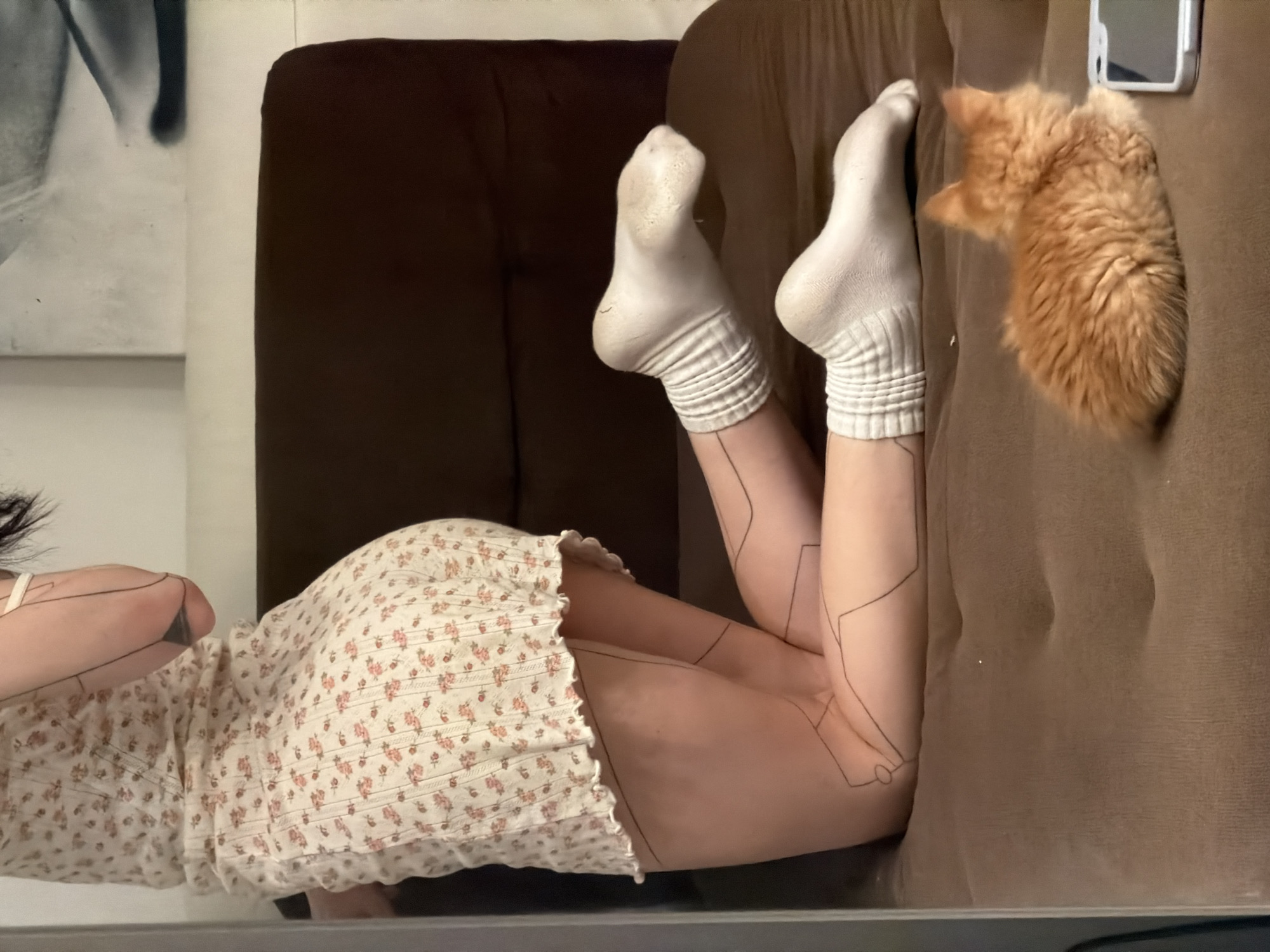 A person wearing a floral dress and white socks is lying on a brown couch with an orange cat next to them. A smartphone is visible on the couch near the cat. A painting is partially visible on the wall. The location in the city of Khimki, Russia cannot be determined from this image.