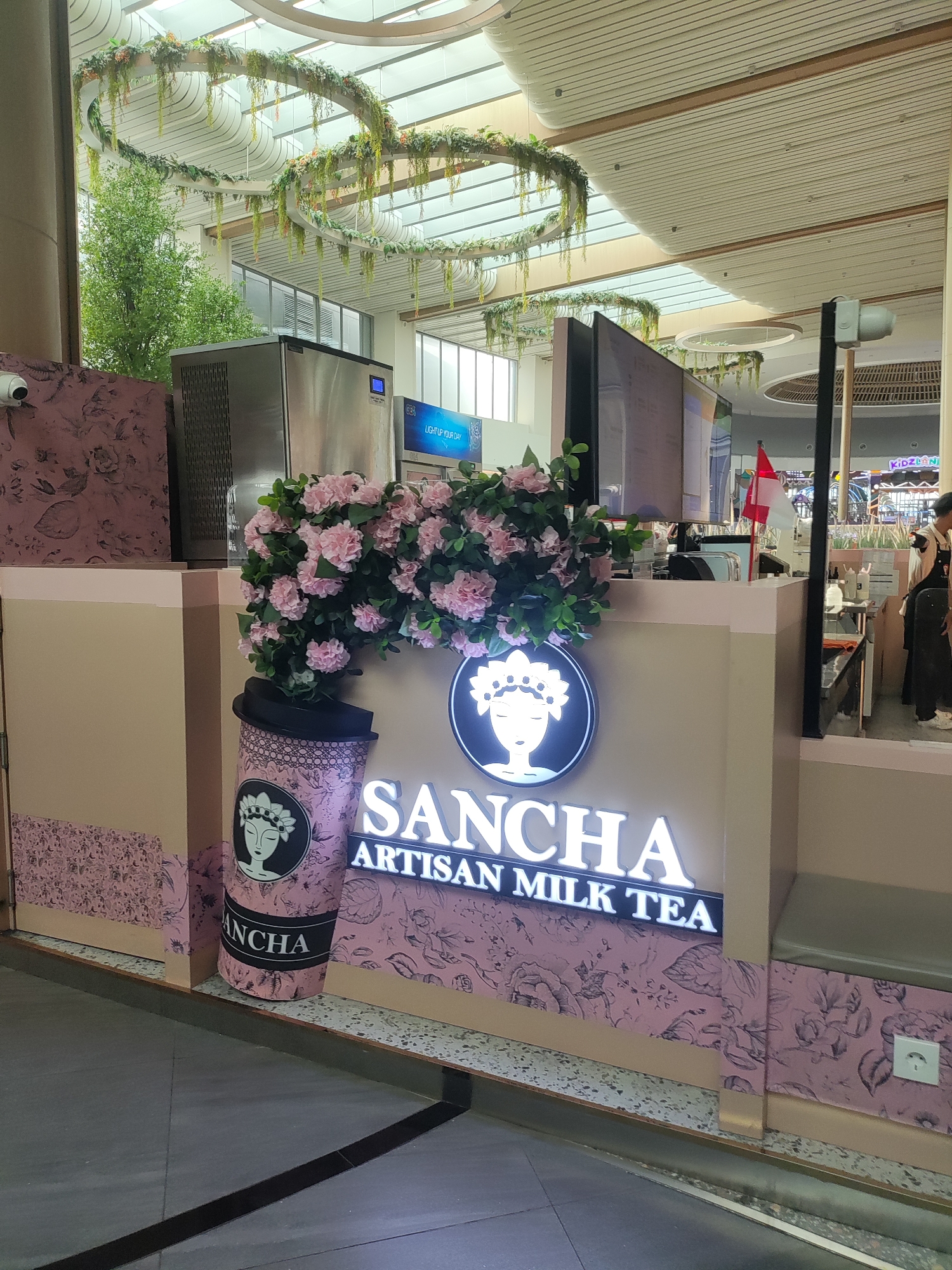 The image shows a San-cha Artisan Milk Tea stall in a bright, modern indoor setting, likely a shopping mall or airport. The stall has a light tan facade decorated with a pink floral pattern. A large, stylized pink cup is placed on the left side, featuring a black and white illustration of a woman's face wearing a crown and the word "SANCHA" in black lettering. Above the cup, a cluster of artificial pink flowers and green leaves is arranged. The main signage for "SANCHA ARTISAN MILK TEA" is illuminated in white LED letters. To the left of the stall, a silver ice machine is visible, and a security camera is mounted on a brown pillar. The ceiling features modern architectural elements with hanging circular floral decorations. In the background, a counter is staffed by a person wearing a dark apron and standing behind a red and white flag. More shops, including one labeled "Kidzland", are visible further in the background. The flooring is a mix of grey tiles and black lines. The overall impression is that of a clean, well-maintained commercial establishment. There is no visual information in the image that specifically places this scene in Lambangjaya, Indonesia.