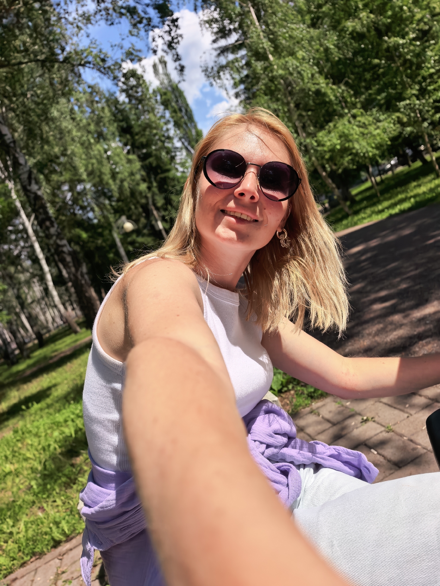 In Sterlitamak, Russia, a blonde woman with sunglasses is captured in a selfie. She is wearing a white tank top and light-colored pants, with a purple scarf tied around her waist. The background shows a park with lush green trees and a paved path. The sunlight filters through the leaves, casting shadows on the ground. The woman is smiling and appears to be enjoying a pleasant day outdoors.