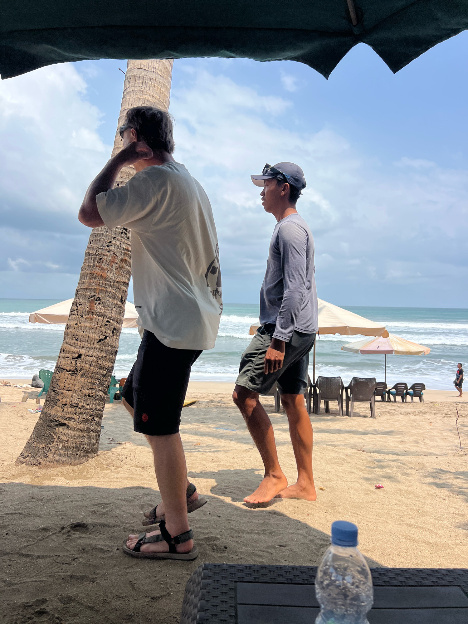 Two men are on a sandy beach in Kuta, Indonesia. The man in the foreground, facing away from the camera, is wearing a white t-shirt, black shorts, and sandals. He is standing next to a palm tree. The second man, facing the ocean, is wearing a grey long-sleeved shirt, dark shorts, and a baseball cap. He is standing with his right foot forward. In the background, the ocean is visible with waves, and beach chairs and umbrellas are set up on the sand. The sky is partly cloudy.