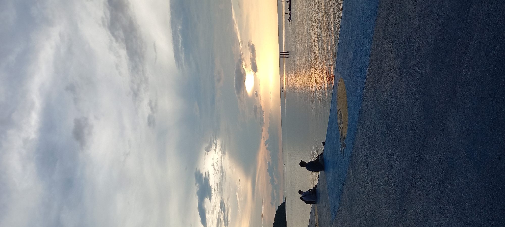 The image captures a sunset scene in Thong Sala, Thailand. The sun, partially obscured by clouds, casts a golden reflection on the water. A concrete surface with a blue and yellow design is visible in the foreground, where two individuals are sitting. In the background, a few structures are seen in the water. The sky is filled with clouds of varying shades.