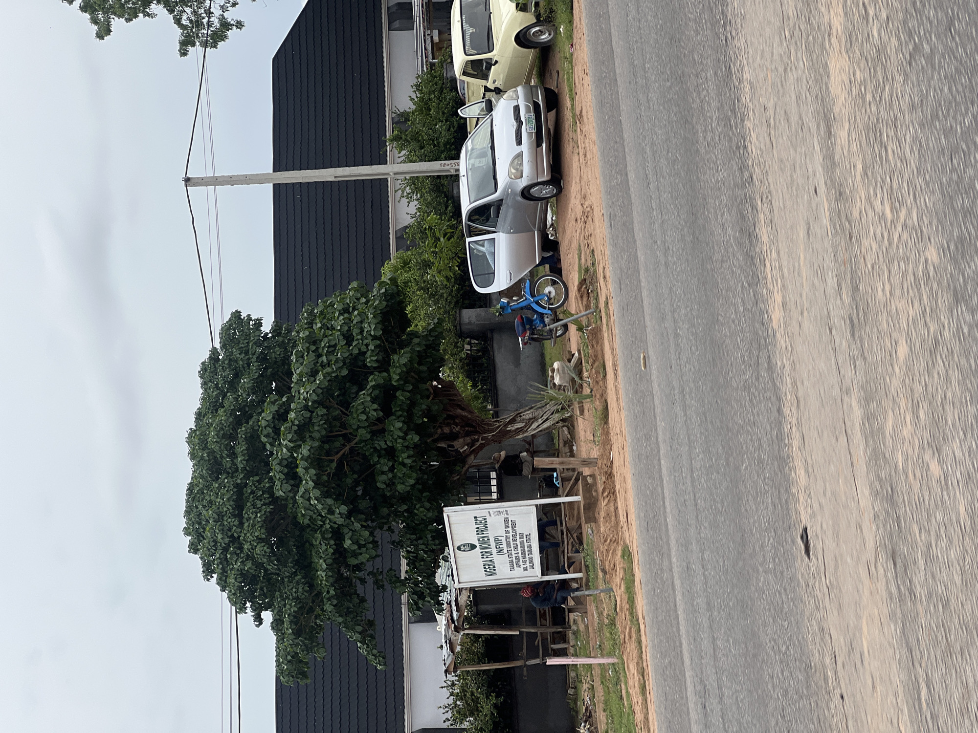 The image captures an outdoor scene on a cloudy day, featuring a road in the foreground and a building with a dark, sloped roof in the background. To the left, a large, leafy tree stands prominently, partially obscuring the building. In front of the tree, a white sign with text is visible, reading "NIGERIA FOR WOMEN PROJECT (NFWP)" and listing "TARABA STATE MINISTRY OF WOMEN AFFAIRS & CHILD DEVELOPMENT" with an address on "HASSARANMA WAY JALINGO, TARABA STATE."
Near the sign, a rudimentary wooden shelter with a corrugated roof is set up. Several people are present, with one individual sitting on a wooden bench under the shelter, appearing to be engaged with their phone. Another person, wearing a red head covering, is seated on a wooden bench near the sign. To the right of the sign, a person is seated on a wooden structure.
Further to the right, on a dirt area adjacent to the road, two vehicles are parked: a silver sedan with its doors open, and a pale yellow older model car. A blue motorcycle is parked between the vehicles and the dirt area. The environment appears to be somewhat unkempt, with loose dirt and some debris scattered around the vehicles and the shelter. The road itself is paved and shows signs of wear. The overall impression is a roadside setting in Jalingo, Nigeria, likely associated with the aforementioned women's project.