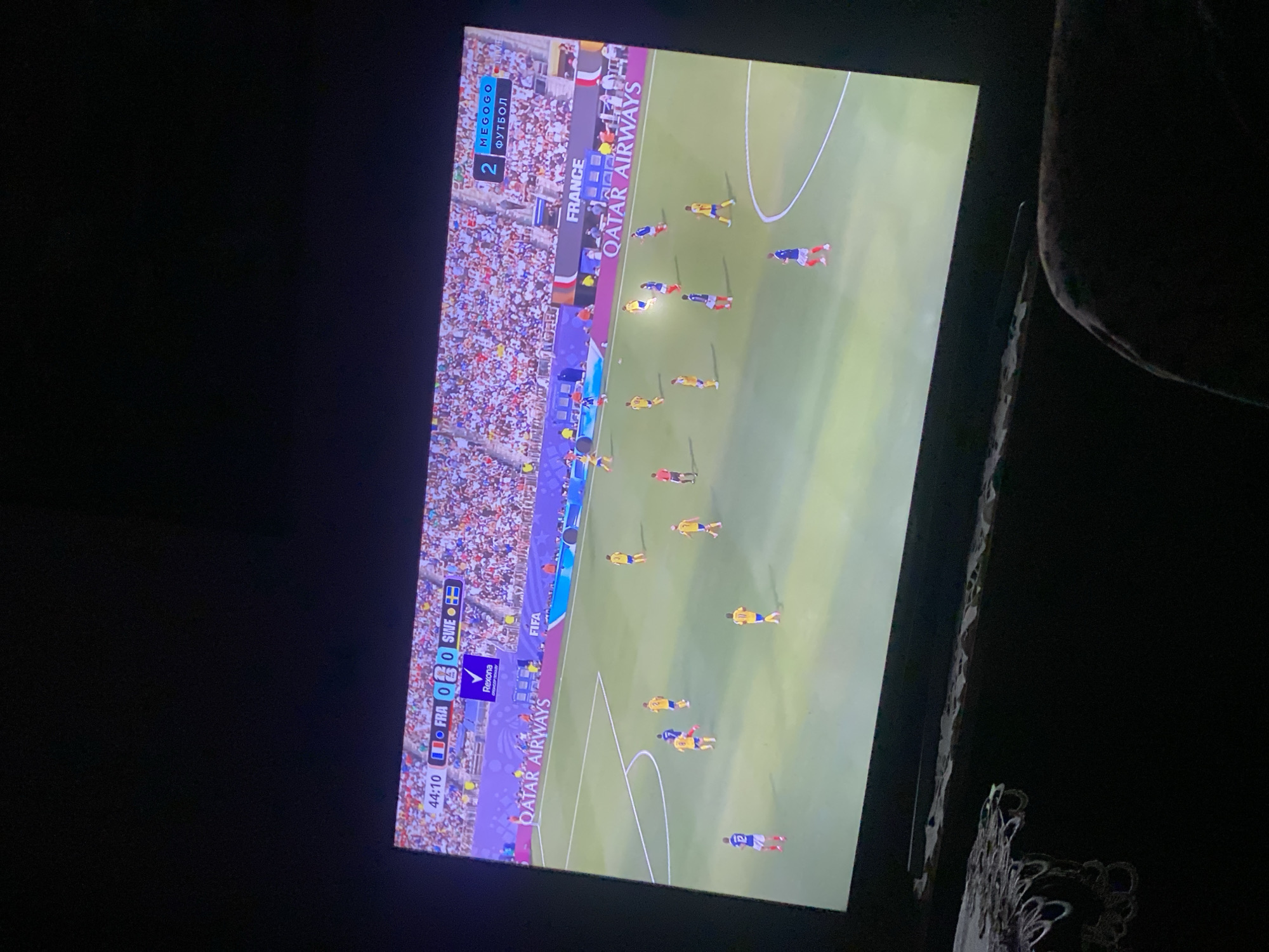 The image shows a television screen displaying a soccer match. The score is 0-0 between France (FRA) and Sweden (SWE) at 44:10. Players in blue and yellow jerseys are on the green field, with a large crowd visible in the stands. Advertising boards for Qatar Airways and FIFA are present around the field. The screen is angled and slightly blurry, suggesting it is being viewed from an off-center position. The environment around the television is dark, with the lower part of the image showing a glimpse of dark furniture and what appears to be a lace doily. This scene is in the city of Zastavna, Ukraine.