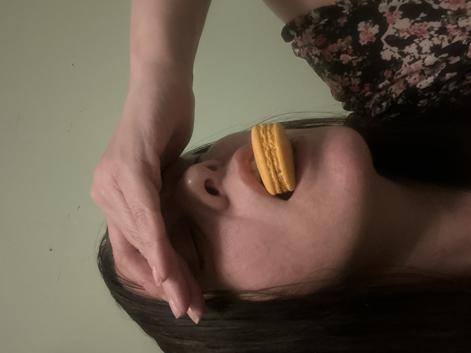 The image shows a woman lying on her back with an orange macaron in her mouth. Her hand is resting on her forehead. She is wearing a floral-print dress. The background is a light green wall. There is no indication of a location within the city of Riga, Latvia, or any specific geographic information.
