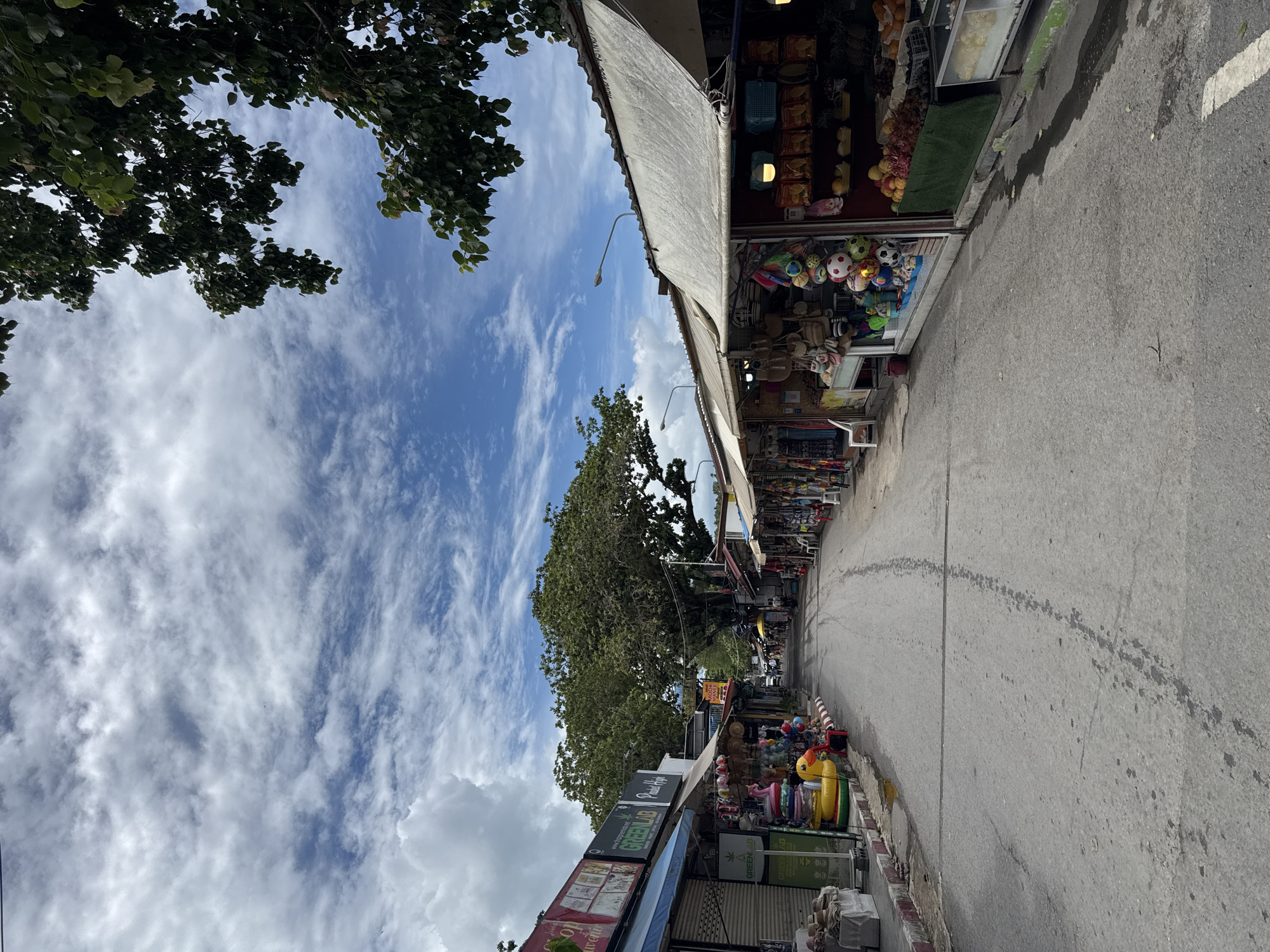 This is a vertical, medium-shot, eye-level, full color photograph of a street lined with shops in Rawai, Thailand. The sky is blue with white, wispy clouds. Trees with lush green leaves hang over the shops. The street is paved with asphalt and appears wet in places. On the left side of the street, shops sell various items, including clothing, inflatable toys (like a yellow duck), and what appears to be souvenirs. A sign for "GREEN LAB" is visible. On the right side of the street, shops display an assortment of goods, including bags, balls, food items, and fruit. One shop has a white canopy, and another has a beige canopy. A few people are visible walking down the street in the distance. Street lights are mounted on poles along the street. The overall scene depicts a bustling marketplace on a cloudy day.
