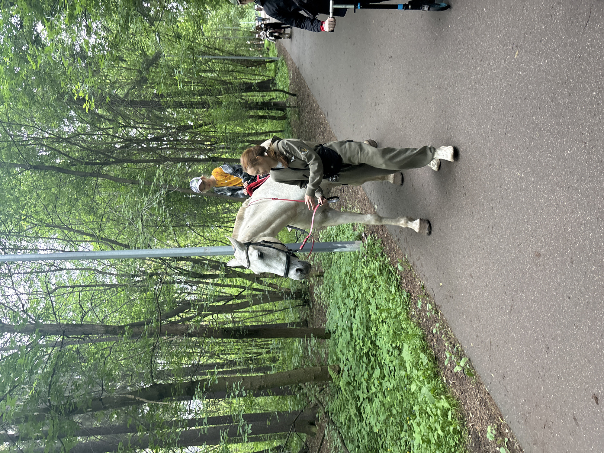 The image displays a recreational scene on a paved asphalt path in a densely wooded park or forest area within Perm, Russia. In the foreground, a white horse, fitted with a black bridle and a red lead rope, is being led left-to-right. A young child with light hair, wearing a yellow long-sleeved shirt, a plaid button-up shirt, and a baseball cap, is riding the horse. A young adult female, with reddish-brown hair pulled back, wearing a khaki-colored utility jumpsuit, white athletic shoes, and a black waist bag, is walking alongside the horse, holding the red lead rope and looking slightly to her right with a faint smile. On the far right, a partially visible individual in a dark jacket is operating a black and blue kick scooter. The path is flanked by abundant green undergrowth and numerous tall trees, primarily deciduous, forming a dense forest canopy. Further down the path, in the background, other individuals, including at least one other person riding a horse, are visible. A tall, slender light-grey pole stands vertically near the horse's head on the left side of the path. The lighting is diffuse, suggesting an overcast day.