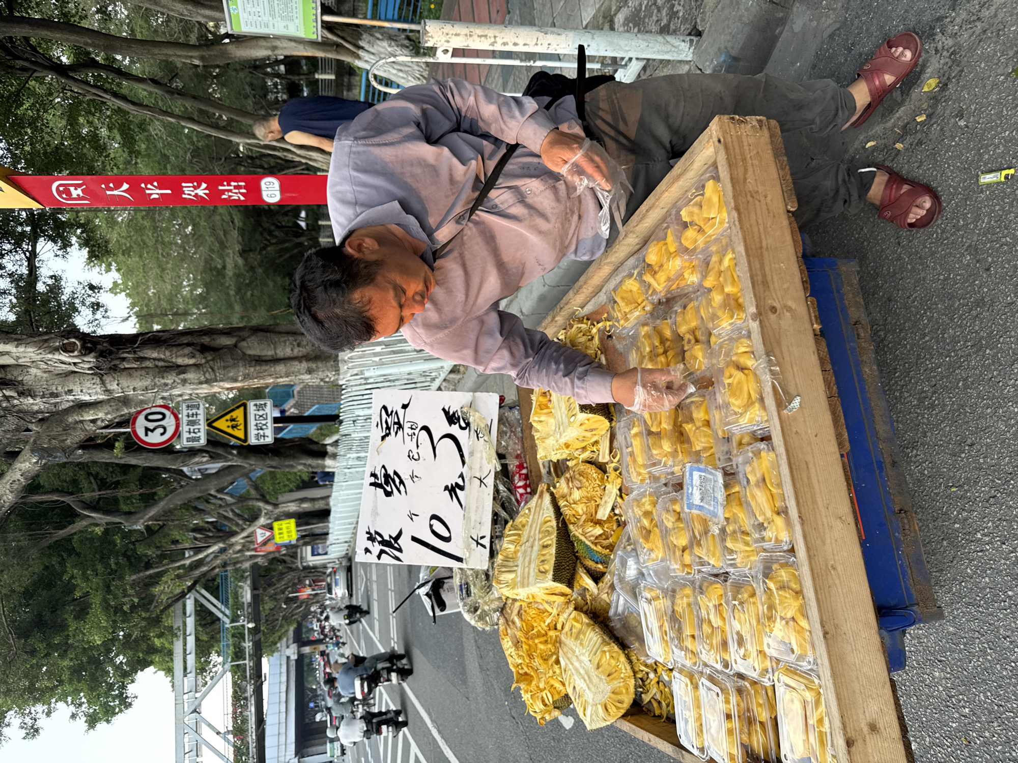 An eye-level, medium-angle shot depicts a vendor selecting jackfruit on a street corner in the Tianhe District of China. The vendor, a middle-aged Asian man with short black hair, wears a light purple button-down shirt and grey trousers. He dons plastic gloves as he carefully picks individual jackfruit pieces from a wooden crate filled with both packaged and unpacked portions. The crates sit on the sidewalk. A handwritten sign advertises "10 yuan for 3" pieces of jackfruit.
In the background, a street scene unfolds with scooters and pedestrians moving along the road. Street signs and tree branches line the road. A red pole displays the Chinese characters "天平架站" and "619", indicating the Tianpingjia Station, a location in the city of Tianhe District.