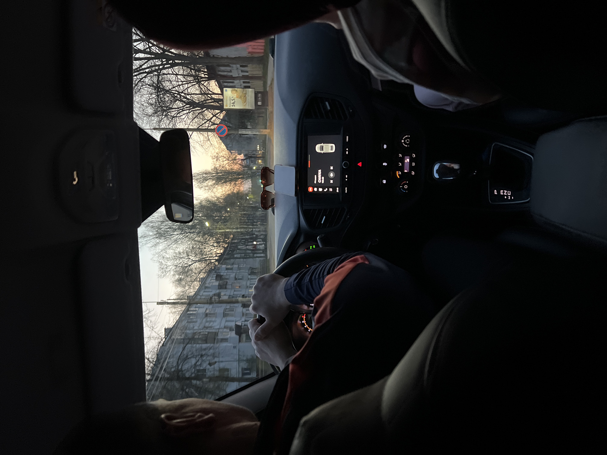 A vehicle with at least two occupants travels on a street in Chaikovsky, Russia. The driver grips the steering wheel, the interior dashboard display visible along with sunglasses and a smartphone. Outside the vehicle, buildings and trees line the street, with a sign displaying "14.5°C" and a circular road sign in view. The angle of the photo gives a high-positioned vantage point inside the car.
