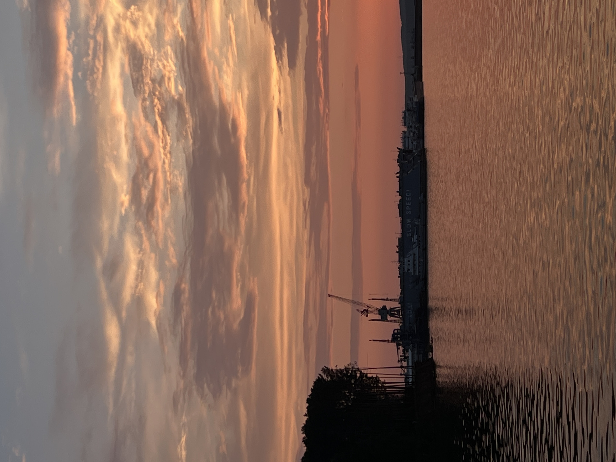 The image depicts a sunset over a body of water, likely the Kama River, in the city of Chaikovsky, Russia. Silhouetted against the vibrant orange and pink sky are a line of trees on the left, followed by a harbor with cranes, ships, and other industrial structures. The water reflects the colors of the sunset, creating ripples of light on its surface. The overall scene is serene and captures the tranquil beauty of a coastal sunset with industrial elements.
