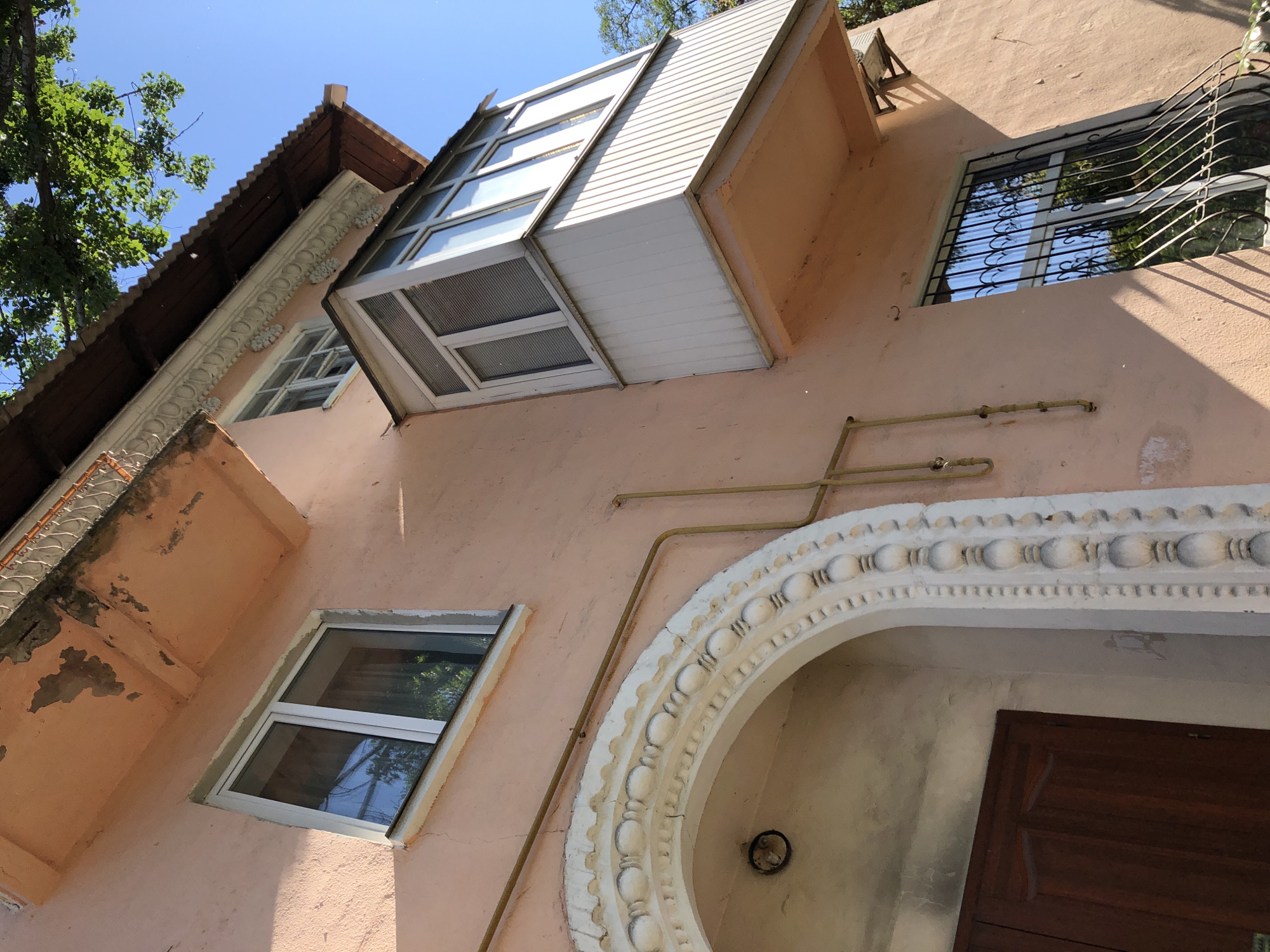 A low-angle, medium shot captures a pale pink, weathered building with an arched entryway on the left, a small balcony jutting out from the center, and a window with wrought-iron bars on the right. The entryway features a dark wooden door and ornate white plasterwork framing the arch, adorned with a pattern of rounded decorative elements. Above the entryway, a window with a white frame reflects the sky and surrounding trees. A yellow pipe runs horizontally across the building's facade, connecting to vertical pipes. The balcony is enclosed with windows and has a pale gray siding. The window on the right also has a white frame and is secured with decorative black wrought-iron bars, reflecting the clear blue sky and green foliage. A small, dark-colored air conditioning unit is mounted on the wall below the roofline to the right of the balcony. The building's stucco exterior shows signs of age, with some chipped paint and cracks. The roof above the balcony is brown and corrugated, extending over the building. Lush green leaves and branches of a tree are visible in the upper right corner, extending into the frame against the bright blue sky. The scene is illuminated by bright sunlight, casting shadows across the building.