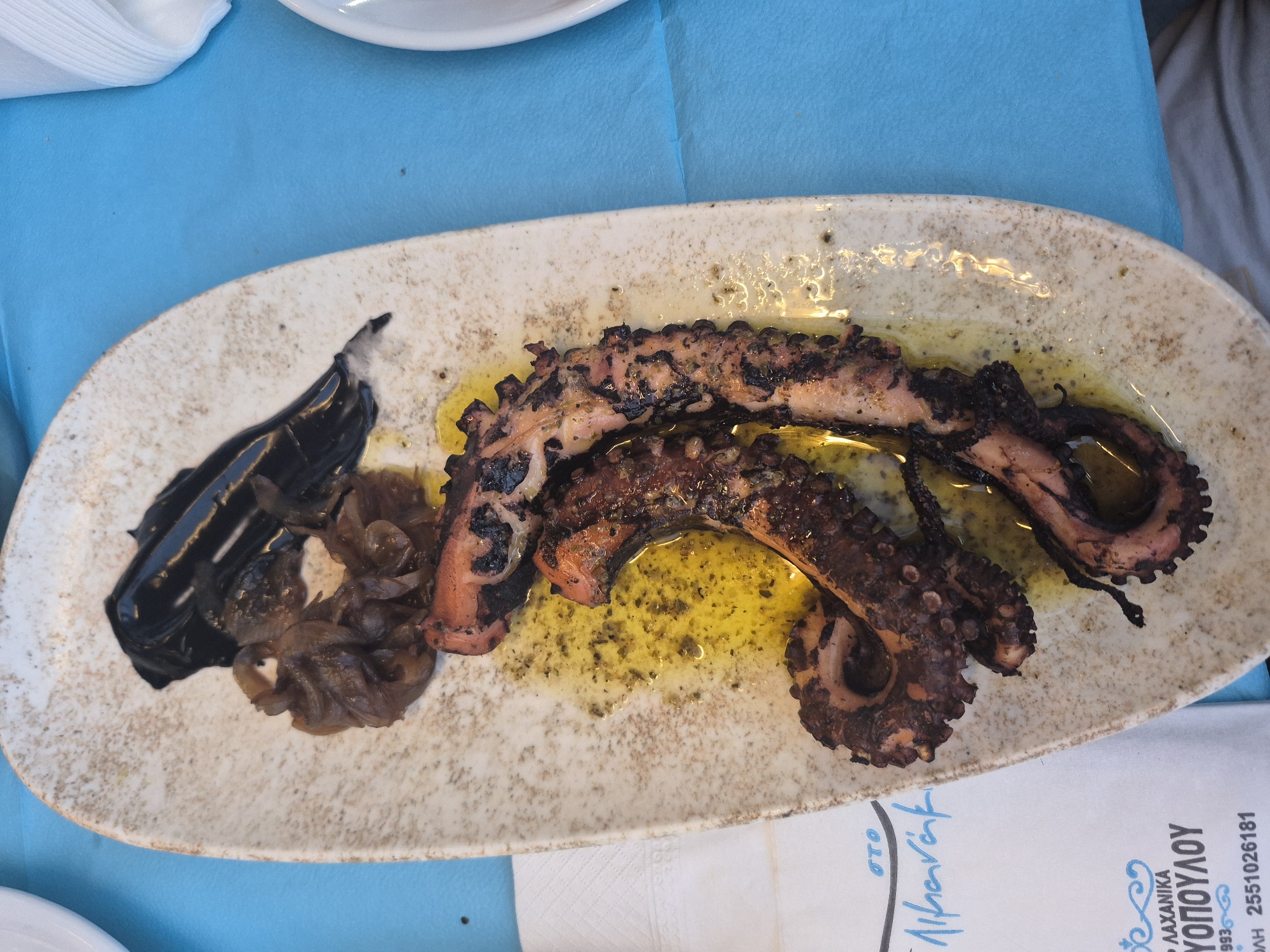 An overhead shot displays a plate of grilled octopus tentacles seasoned with herbs and oil, accompanied by caramelized onions and a dark, gelatinous sauce, possibly squid ink, presented on a light-colored, speckled ceramic plate. The plate rests on a vibrant blue tablecloth, with a white napkin visible to the left and a white plate edge to the right, suggesting an outdoor dining setting. A partially visible paper menu in the lower left corner bears Greek text and the number "2551026181", indicating the location is likely a restaurant in Makri, Greece, a coastal town known for its seafood. No people are visible in the frame.