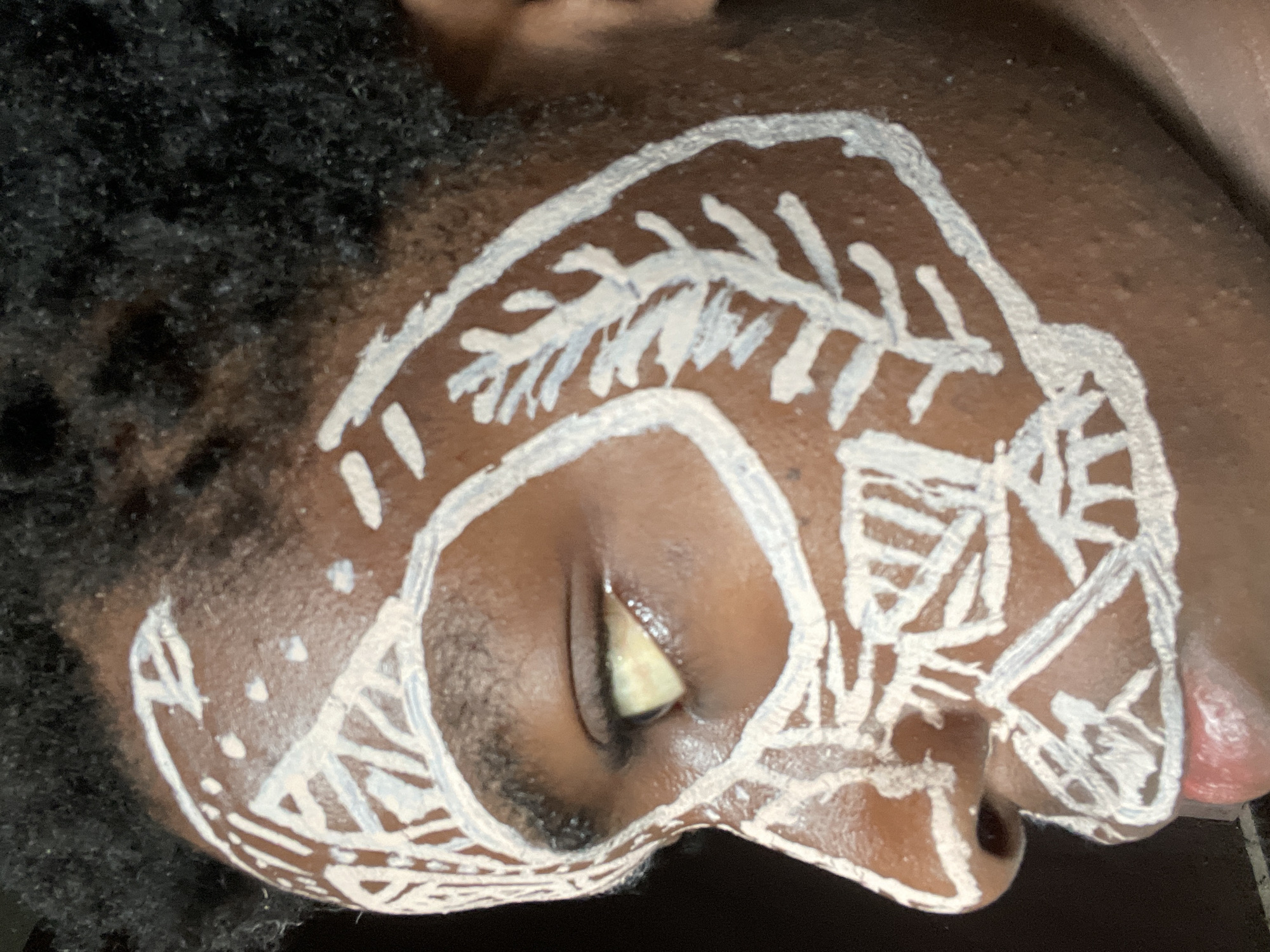 A close-up photograph depicts a person's face, specifically the forehead, eye, nose, and cheek, with white tribal-style paint markings. The person has dark skin and tightly curled black hair is visible on the left side of the frame. The eye is partially closed. The background is dark and indistinct. The location in the city of Nkpor, Nigeria is not visually evident in the image.
