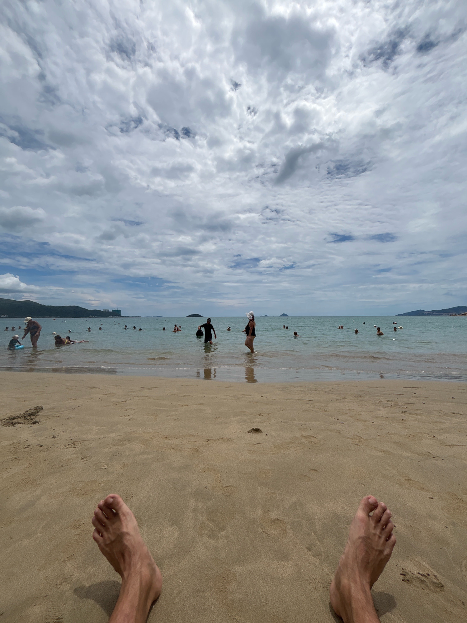 The image depicts a wide shot of a crowded beach in Nha Trang, Vietnam. The foreground is dominated by the sandy beach with two bare feet positioned at the bottom of the frame. In the mid-ground, the ocean is filled with numerous people swimming, wading, and standing in the water. Some individuals are further out, appearing to be enjoying a swim, while others are closer to the shore. The water is a light blue-green color and appears calm. In the background, a hazy coastline with rolling hills and a few buildings can be seen. The sky is filled with dramatic, white, and gray clouds. The overall scene conveys a busy, sunny day at the beach with many people engaged in water activities.