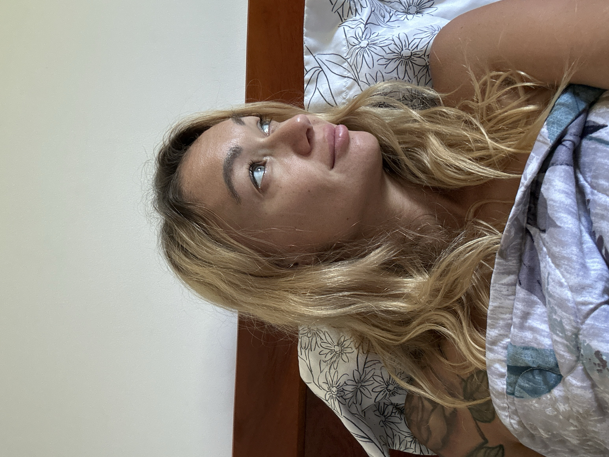 A medium shot shows a blonde woman lying in bed, looking to her right. She has fair skin, freckles, blue eyes, and blonde, wavy hair that falls over her shoulders. She is wearing a floral-patterned top. A patterned duvet covers her lower body. The background consists of a white wall and a wooden headboard. The scene is indoors, likely a bedroom in Mũi Né, Vietnam.
