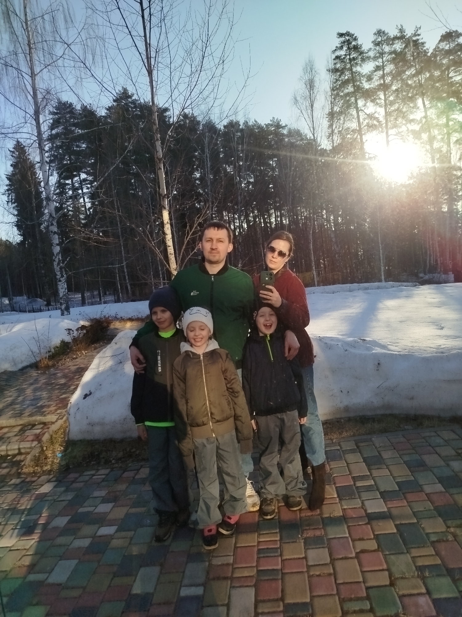 A photograph depicts a group of five individuals—two adults and three children—posed outdoors. The adults stand in the back, with a man in a green jacket on the left, his arm around the shoulder of the child on his left. A woman, wearing sunglasses and a burgundy top, stands to the right, seemingly holding a phone. The children stand in front; two boys flank a girl. A layer of snow covers the ground behind the group, indicating a cold climate or time of year. A forest of trees provides a backdrop, with the sun shining through them. The group stands on a paved area, which appears to be constructed from interlocking brick or stone. The scene seems to be a snapshot taken in a suburban or rural area with a residential feel, possibly a park or recreational area. Given the information about the city of Izhevsk, Russia, it suggests a setting within or around the outskirts of this location.