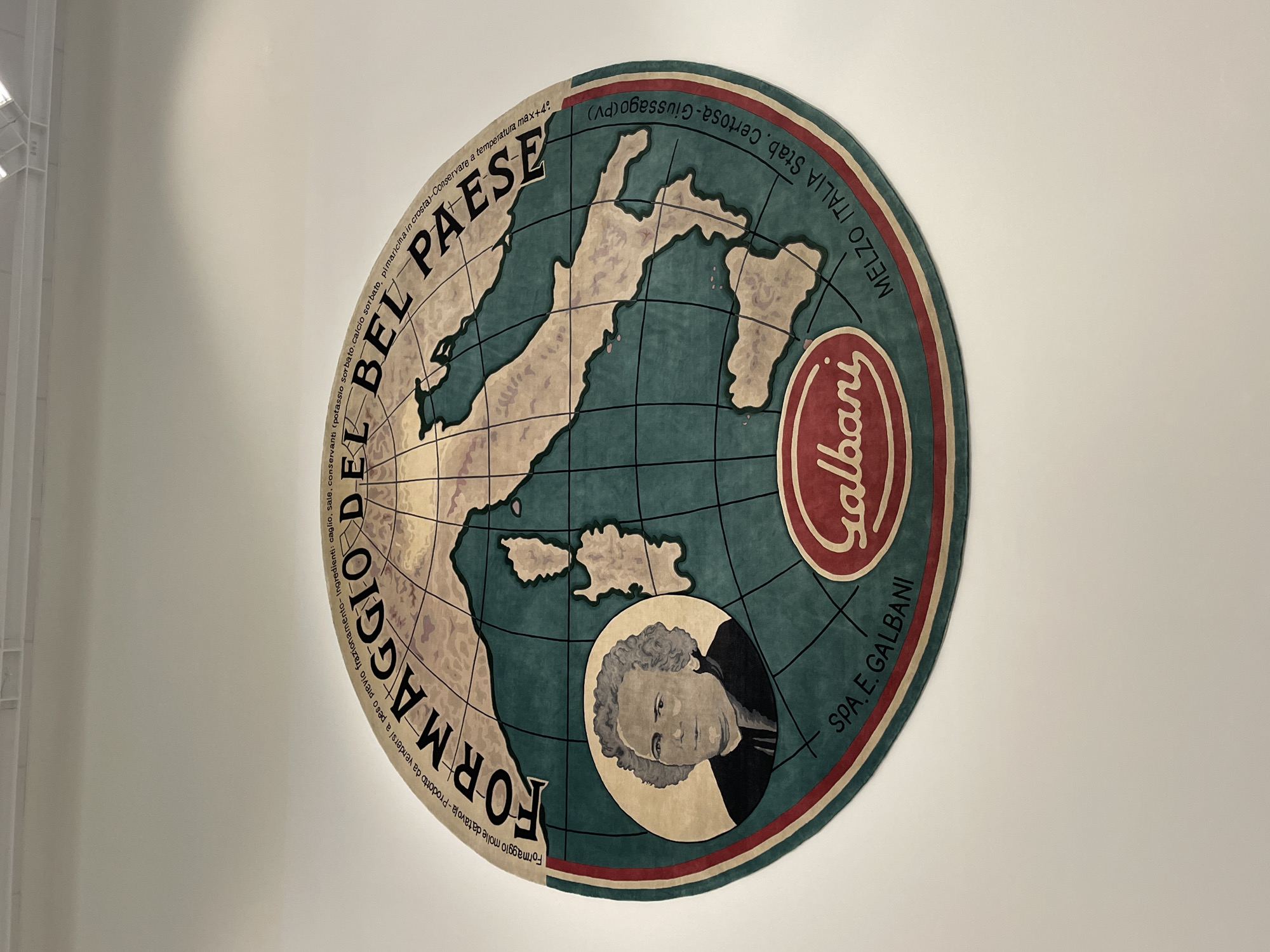 The image shows a round, promotional sign or display, likely made of lightweight material and mounted on a plain white wall. The design is an artistic representation of a world map, primarily focused on Italy, with the map being green and land areas being beige. Text reading "FORMAGGIO DEL BEL PAESE" is printed along the lower left perimeter. The sign is printed with the word "Galbani" in cursive letters within a red and tan oval near the right edge. Under the map, there is a circular portrait of a man. The sign also includes the text "MELZO ITALIA Stab Certosa-Giussago (PV)". More text reading "Formaggio molle datavola-Prodotto da vendersi a peso previo frazionamento-Ingredienti: caglio, sale, conservanti (potassio sorbato.calcio sorbato, pimaricina in crosta)-Conservare a temperatura max +4°" and "SPA. E. GALBANI" can also be seen on the sign. The image itself suggests that the item is currently located indoors on a wall. There is nothing in the image that indicates that this image was taken in Metz, France.
