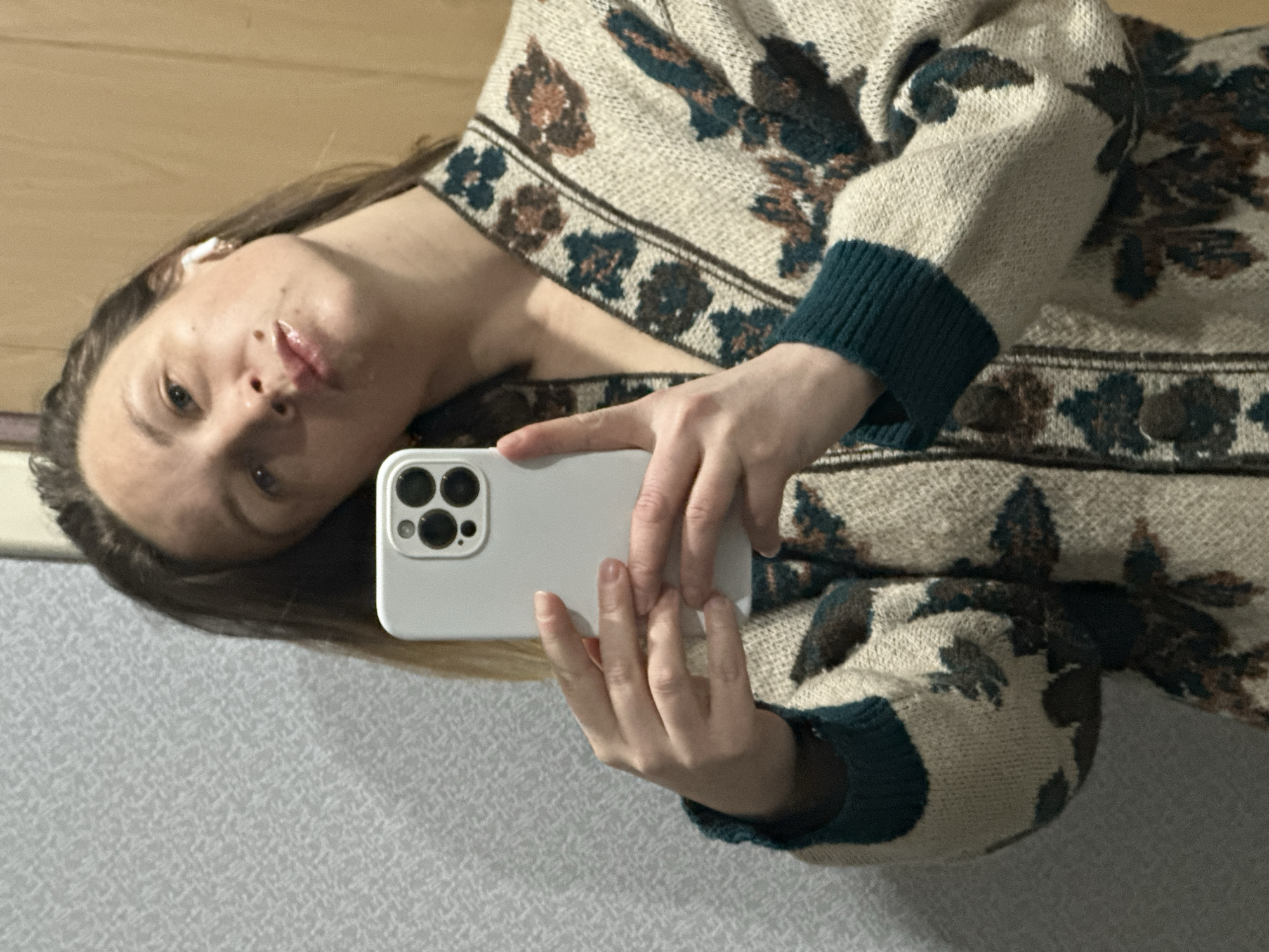 A young woman is depicted taking a mirror selfie indoors. The image is oriented sideways, showing her head tilted to the right. She has long brown hair and a neutral expression, gazing directly at the camera with slightly parted, glossy lips. A white earbud is visible in her left ear. She is wearing a knitted cardigan or sweater with a light beige base, adorned with a repeating pattern of stylized floral or organic motifs in dark green and brown. The cuffs of the sweater are a solid dark green. In her right hand, she holds a white smartphone, identifiable by its triple-camera setup and flash module, likely an iPhone, positioned to capture the reflection. The background includes a plain, light brown wall behind her head and a textured, light gray surface below, consistent with an indoor setting. The lighting appears to be artificial. There is no visible text in the image. The general location context is Chaikovsky, Russia.