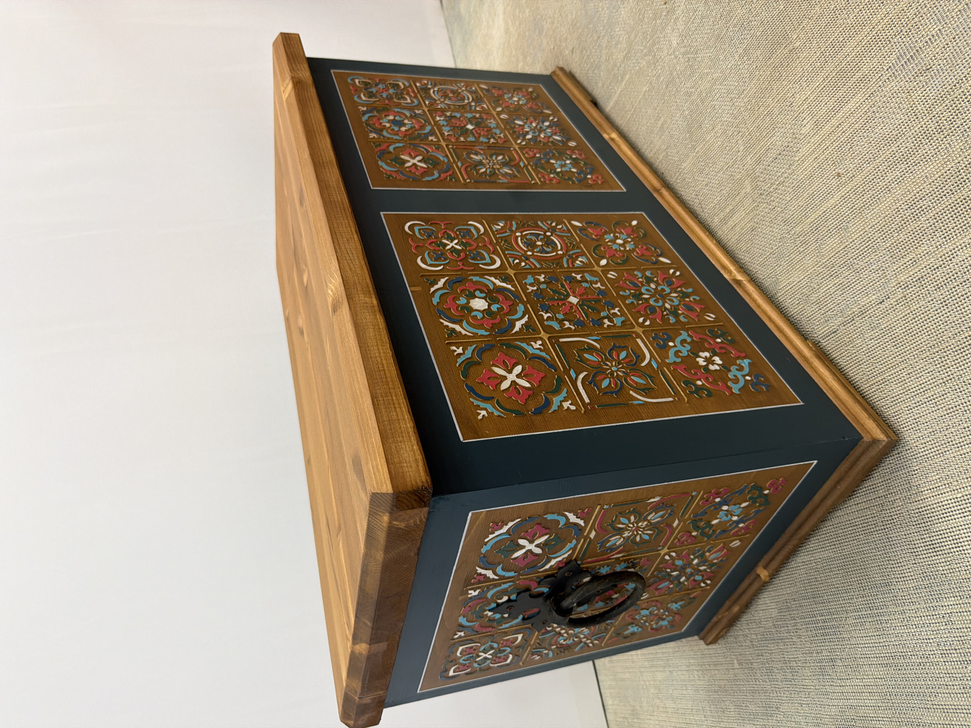 The image shows a decorative wooden chest with a natural wood lid. The sides of the chest are painted a dark teal, and feature panels decorated with a mosaic of colorful, intricate tile-like patterns in shades of red, blue, white, and brown. A dark metal ring handle is attached to the front left side of the chest. The chest is placed on a textured, light-colored surface, possibly a rug or carpet. The background is a plain white wall, suggesting an indoor setting. There is no indication of the city of Maloyaroslavets, Russia, or any other specific location.