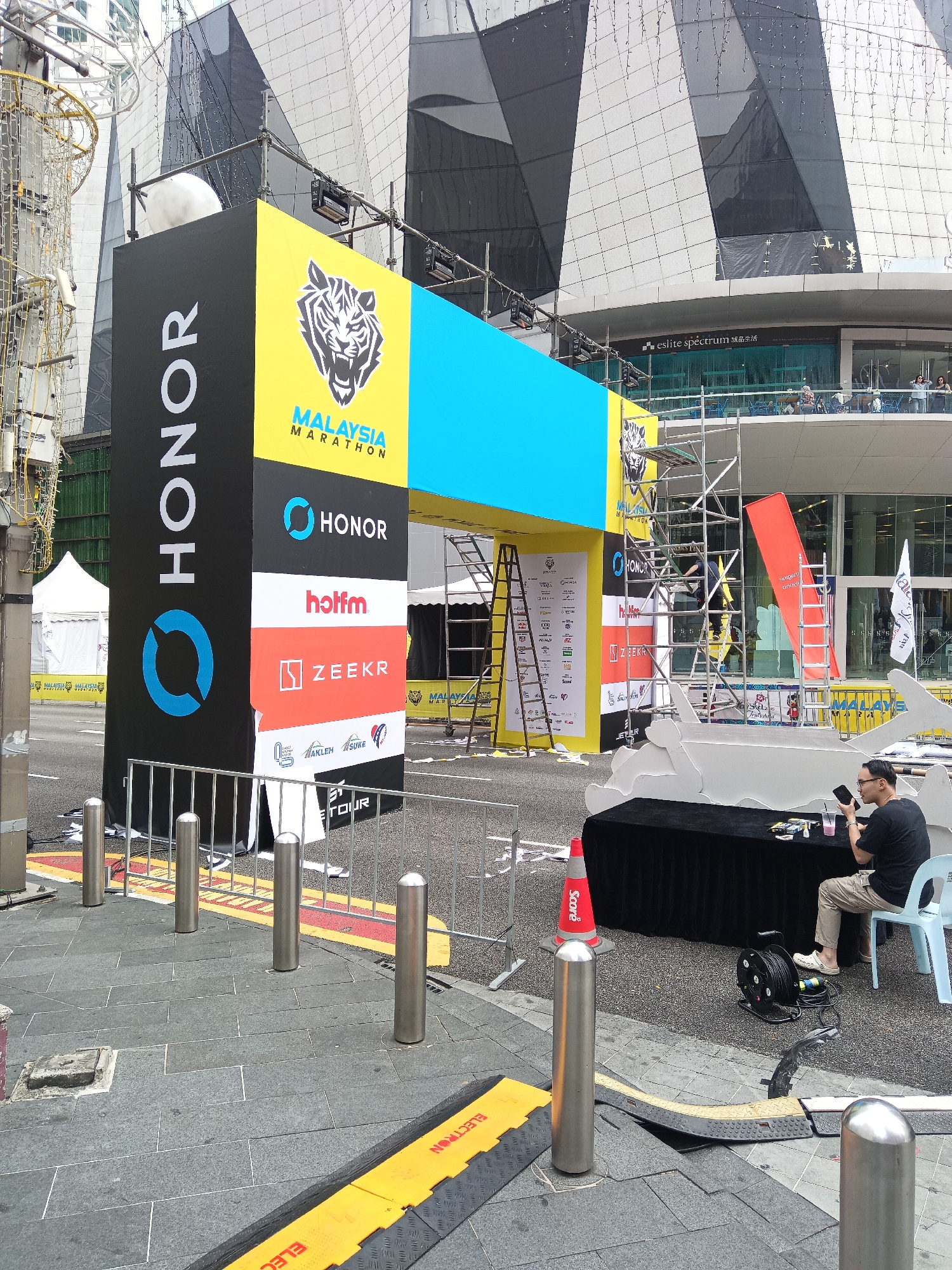 This image captures a section of the Malaysia Marathon entrance arch and its immediate surroundings, likely located in the heart of Kuala Lumpur's central business district, given the modern skyscraper architecture in the background. The arch is a large, multi-sided structure featuring prominent branding for "HONOR" on a black panel and "MALAYSIA MARATHON" with a tiger graphic on a yellow panel. Other sponsor logos, including "hotfm" and "ZEEKR," are visible on vertical panels.
Scaffolding is erected around the arch, suggesting ongoing setup or maintenance. A red ladder leans against the scaffolding. In the background, a large, modern building with a distinctive geometric facade dominates the skyline. Several tents are set up behind the arch, likely for event support or registration.
In the foreground, a man in a black t-shirt and light-colored pants sits at a black draped table, holding a smartphone and appearing to be engaged with it. A light blue plastic chair is beside him. Various items, including what appear to be drinks and small boxes, are on the table. Electrical cables and a cord reel are on the ground near the table. A red traffic cone with the word "Score" is positioned near the table. The ground is paved with dark gray tiles, with some yellow and black hazard tape visible along the edges of the roadway. Several silver bollards are placed along a metal barrier. A large, white, cut-out shark-like structure is also visible in the scene, positioned near the table and the man.