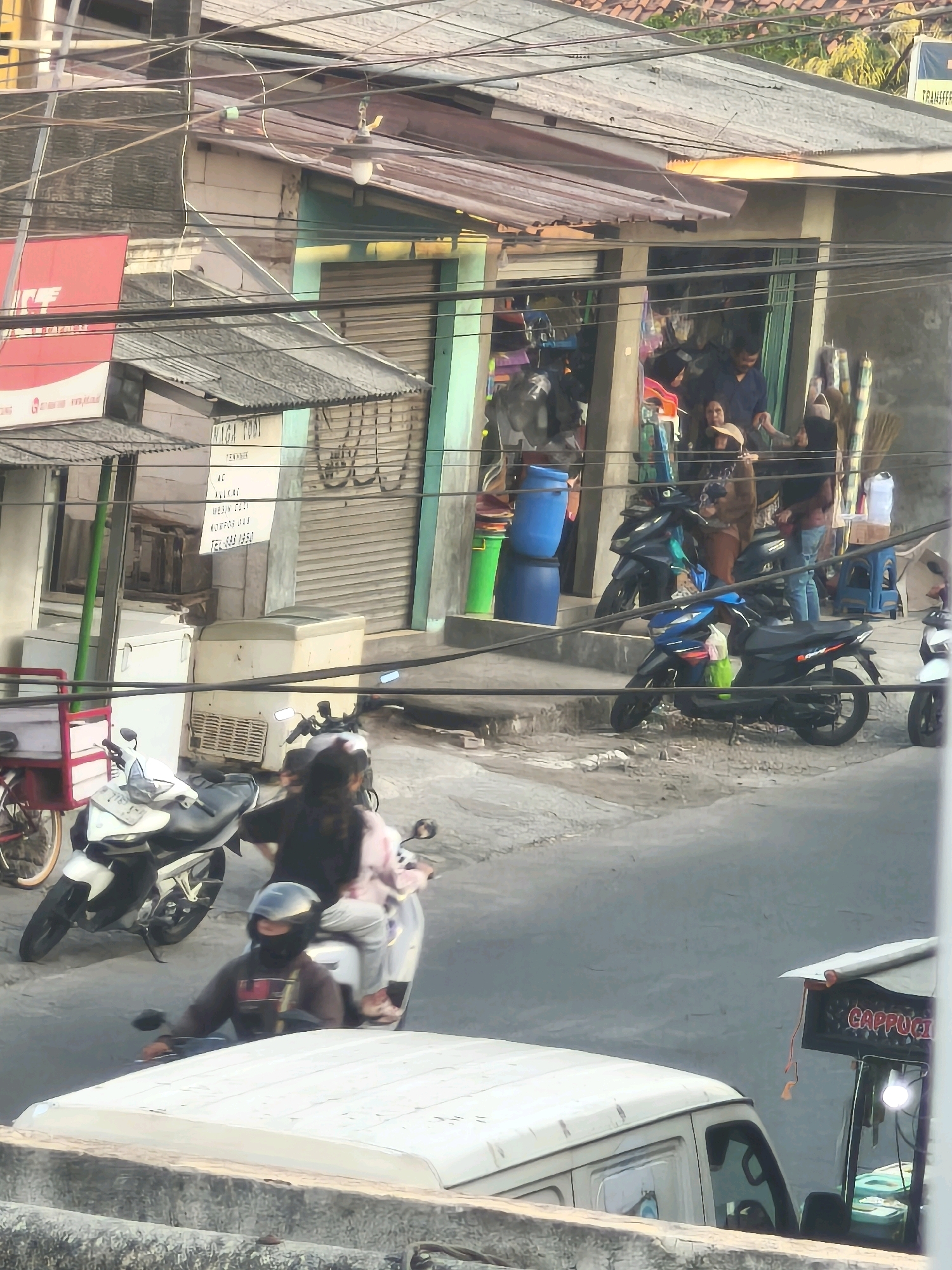 The image captures a street scene in an urban environment, possibly in Southeast Asia. Several motorcycles are parked and in motion. In the foreground, a white van with a light-colored roof is partially visible. Two people are riding on a motorcycle in the middle ground. The driver wears a helmet and a dark-colored jacket, with a black mask covering their lower face. The passenger, wearing a pink top, is seated behind the driver. Another white motorcycle is parked to the left of this pair. Behind the motorcycles, a shop with an open entrance displays various items, and several people are visible inside and near the entrance. One person is seen holding a straw hat. A tall blue barrel is positioned outside the shop. Further back, another dark-colored motorcycle is parked. The street is paved, and overhead power lines crisscross the scene. Buildings with corrugated metal roofs and shuttered windows line the street. A small cart with a sign reading "CAPPUCI" is visible on the right side of the image. The overall atmosphere suggests a busy marketplace or commercial area.