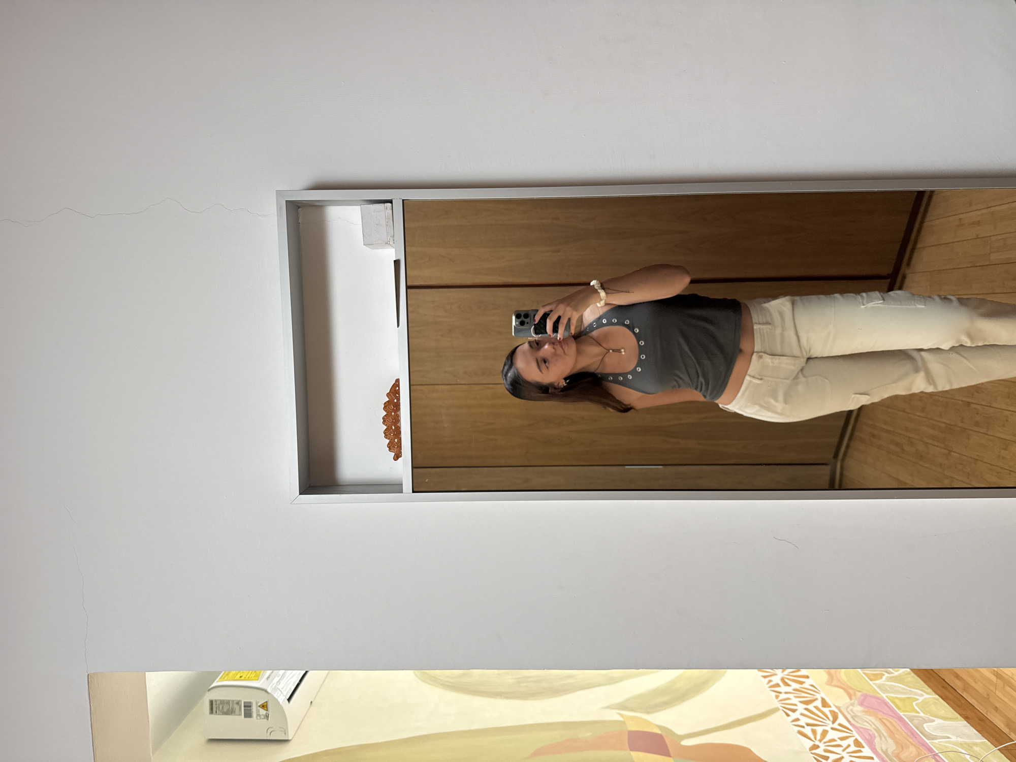 A woman is taking a selfie in a mirror. The mirror is mounted on a white wall with cracks. In the background of the reflection, there is a wooden cabinet and a light-colored wooden floor. Below the mirror, there is a painting with curvilinear shapes. The location appears to be inside a building in San Bernardino Tlaxcalancingo, Mexico.