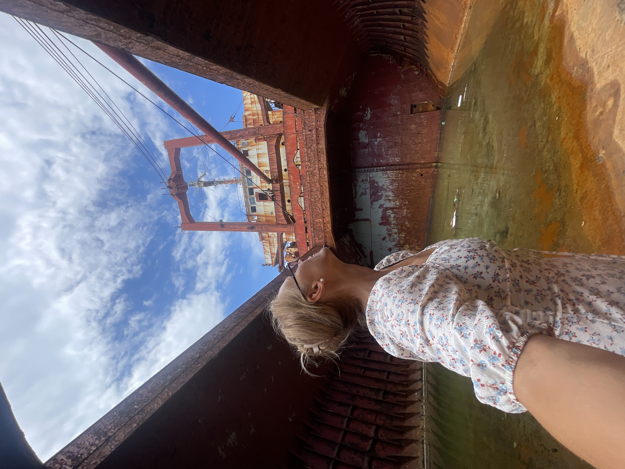 The image depicts the interior of a disused cargo ship, possibly in a dockyard or beached. The ship's hull is heavily rusted, with peeling red paint. The water in the hull is murky and reflects the sky. A woman with blonde hair, wearing glasses and a floral dress, stands in the foreground, looking up at the sky and the ship's structure. The background shows a bright blue sky with scattered clouds, and the upper portion of the ship's superstructure, also showing signs of rust and disrepair. The scene conveys a sense of decay and abandonment. This location is believed to be near the city of Kabardinka, Russia, where several ships have been deliberately grounded to serve as attractions.