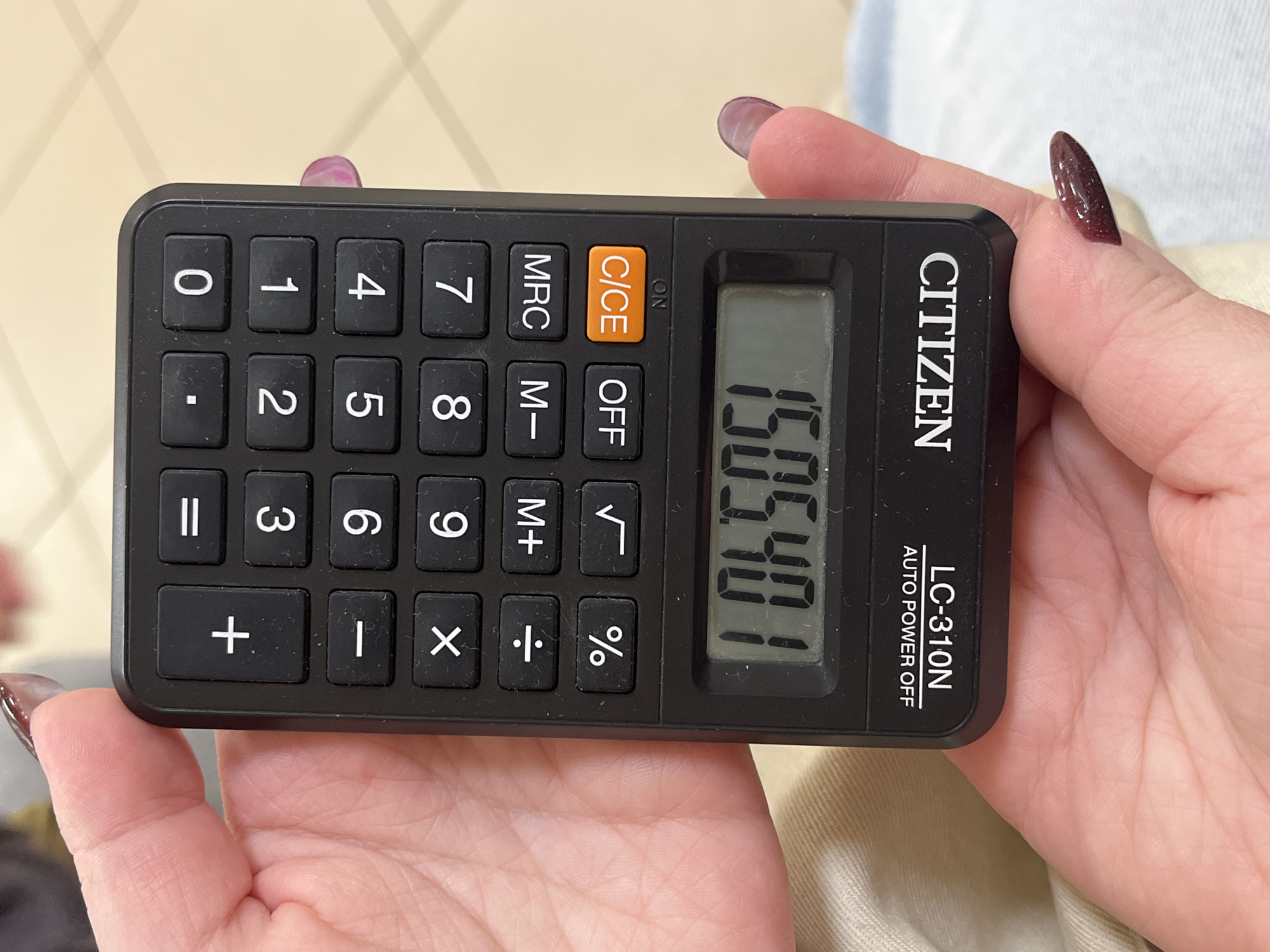 A person is holding a black Citizen LC-310N calculator. The calculator displays the number "1505.401" on the screen. The buttons on the calculator are black with white text, except for the C/CE button, which is orange. The background consists of the person's hand and clothing.