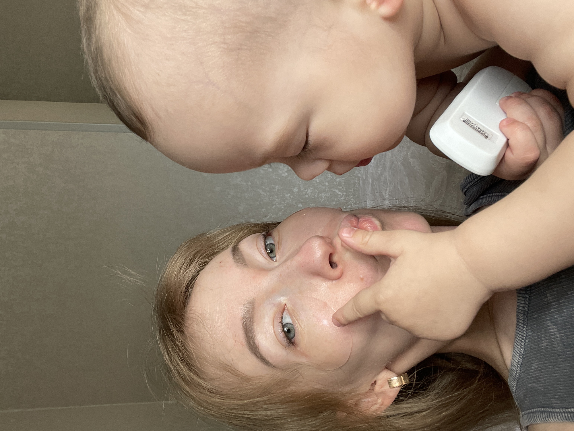A baby holds a white, rectangular device with a sticker on it in its right hand. The baby's head is close to the face of a fair-skinned woman with light-colored hair. The woman is holding her mouth open with her finger. The background is a plain wall. This photo was taken in Perm, Russia.