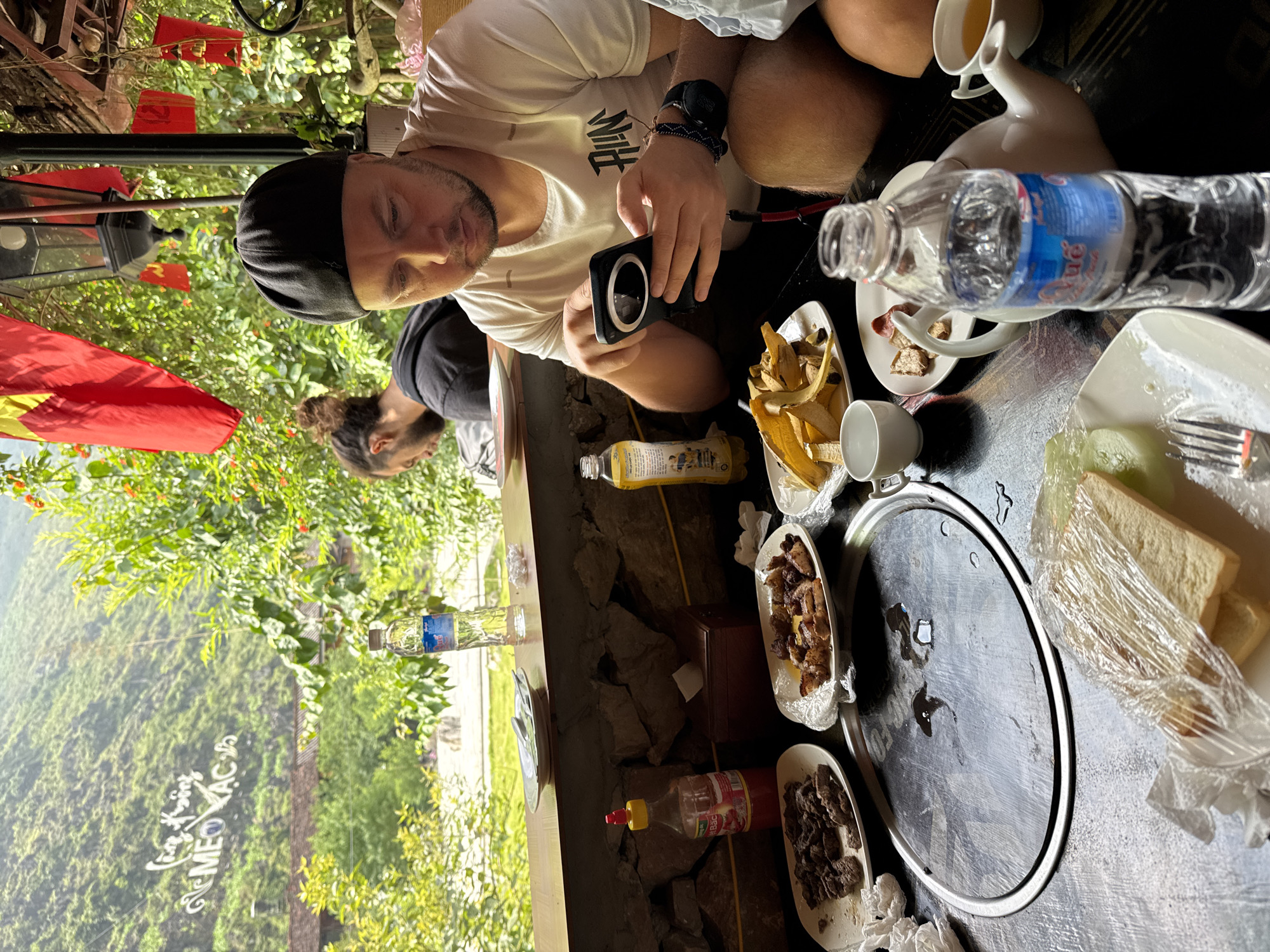 The image shows a man in a white shirt and black cap seated at a table outdoors, holding a black electronic device. A second man with a man bun is visible in the background, slightly out of focus. The table is laden with food, including plates of meat, fries, bread, and various bottles and containers. A circular cooking surface sits in the center of the table. The scene appears to be in a mountainous area, identifiable as Mèo Vạc Commune, Vietnam, based on the distinctive landscape visible through the open-air structure. A Vietnamese flag is also visible in the background.