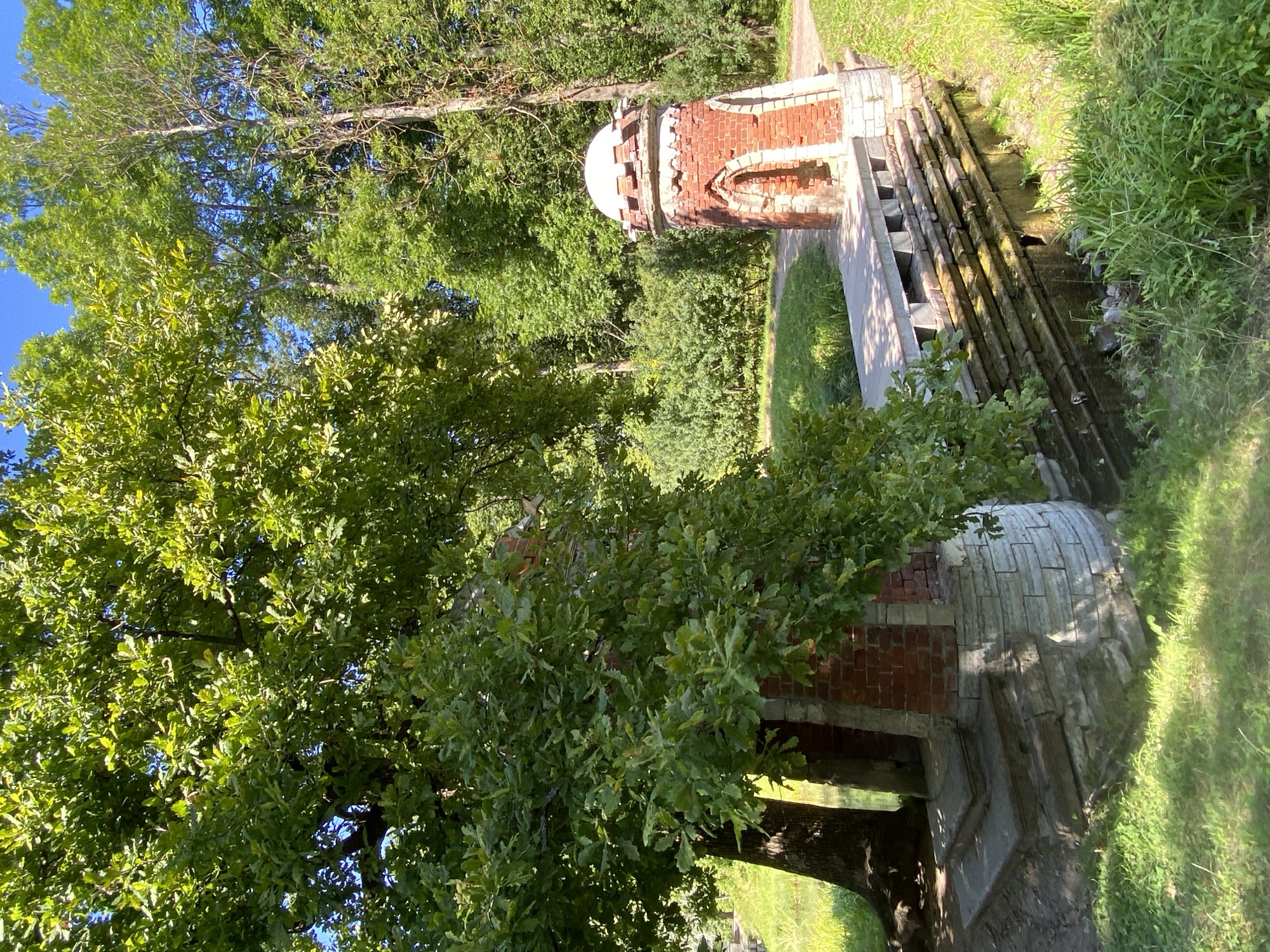 The image shows a bridge in the Catherine Park in Pushkin, Russia. The bridge is made of stone and brick and has a picturesque design with two ornate towers. The towers are red brick with white stone details and topped with small domes. A large oak tree with lush green leaves is on the left side of the bridge, partially obscuring one of the towers. The bridge spans a small waterway. The surrounding environment is a park with green grass, bushes, and trees under a clear blue sky, suggesting a sunny day. No people are visible in the image.