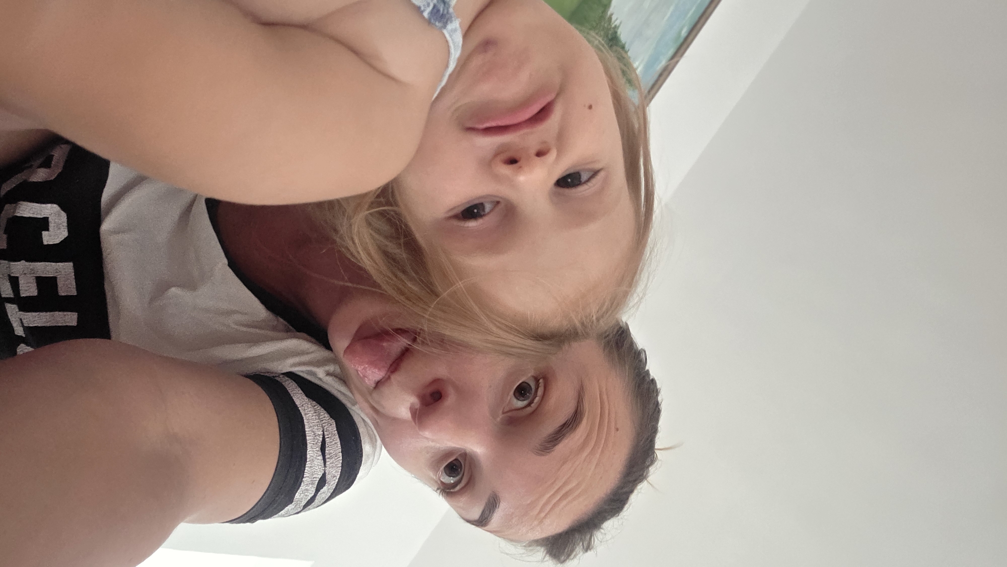 The media file shows a close-up, upside-down selfie of two people, likely a mother and child, in an indoor setting. The person in the foreground is an adult woman with dark hair, brown eyes, and prominent forehead wrinkles. She is wearing a white t-shirt with black stripes on the sleeves and what appears to be text on the front that is partially visible, reading "CEL". She is sticking her tongue out, with a playful expression.
Behind her, and slightly above, is a young child with blonde hair and brown eyes. The child is looking towards the camera with a curious expression and has their mouth slightly open. They are wearing a blue and white patterned garment. The background is a plain, light-colored wall, suggesting an indoor environment. A portion of what looks like a framed artwork with a green and blue landscape can be seen in the upper right corner, above the child's head. The lighting appears to be bright, indicating it could be daytime. The overall mood of the image is lighthearted and candid, capturing a moment of playful interaction between the two subjects. The location context provided is Guía de Isora, Spain, which does not offer further visual cues in the image itself.
