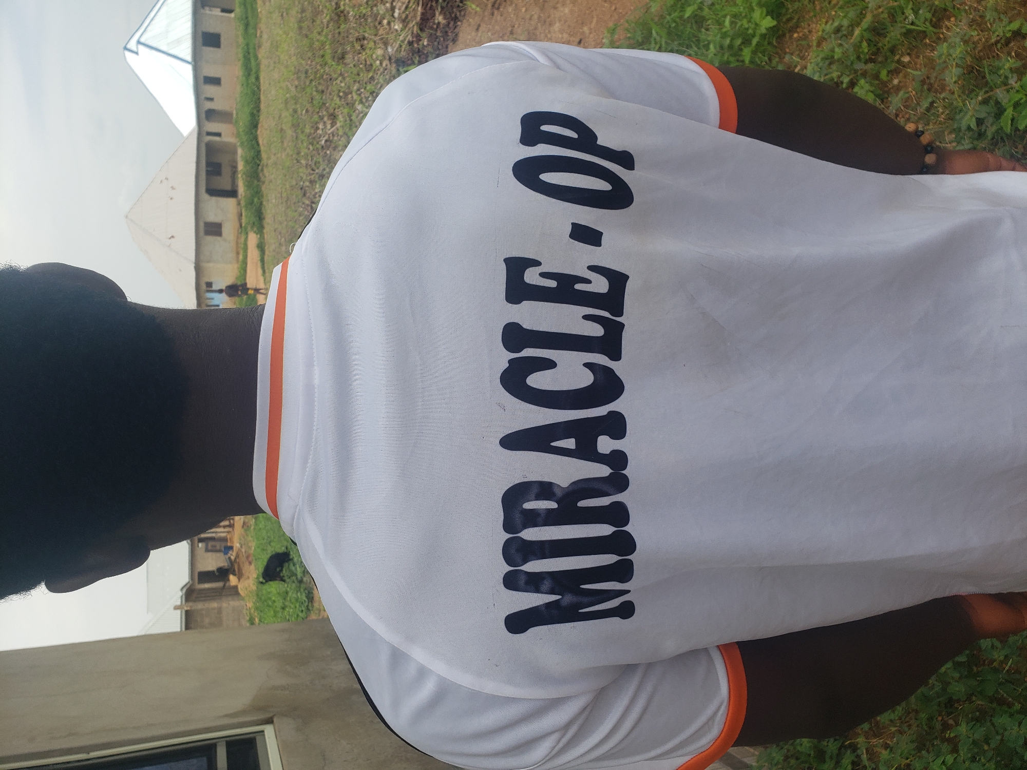 A close-up, rear-facing shot shows the back of a person wearing a white t-shirt with "MIRACLE - OP" printed in dark blue lettering. The t-shirt has orange trim around the collar and sleeves. The background suggests an outdoor environment, possibly a rural or semi-urban area, with grassy terrain and buildings under construction. In the distance, two unfinished structures with distinct rooflines are visible. One building has a grey, pitched roof, while the other features a white, peaked roof. A dark-colored animal, possibly a goat, is present in the middle ground, near the buildings. A person is also visible in the far distance, standing near the construction site. The overall scene appears to be in Kachia, Nigeria, given the architectural style and vegetation.