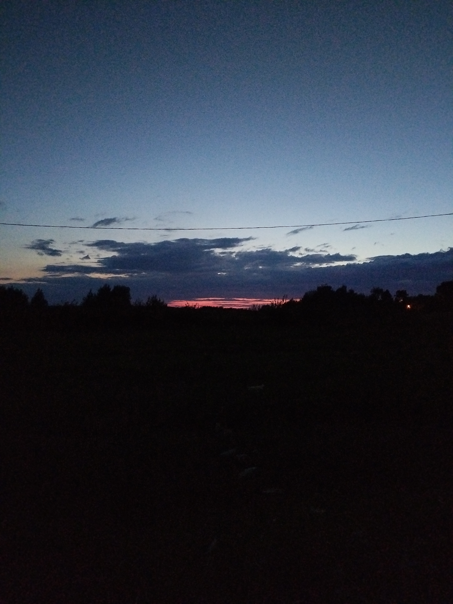 The image depicts a dark, silhouetted landscape at dusk. A thin, horizontal line, likely a power line, is visible across the upper middle section of the frame. Below this line, a band of clouds stretches across the horizon. The sky above is a gradient of deep blue to a lighter blue. The lower half of the image is almost entirely black, obscuring any details of the ground or vegetation. A faint reddish glow is discernible near the horizon line, suggesting the last remnants of sunset. No people, animals, or specific structures are clearly identifiable due to the low light conditions and the predominant darkness. The scene appears to be an outdoor, rural or semi-rural environment.