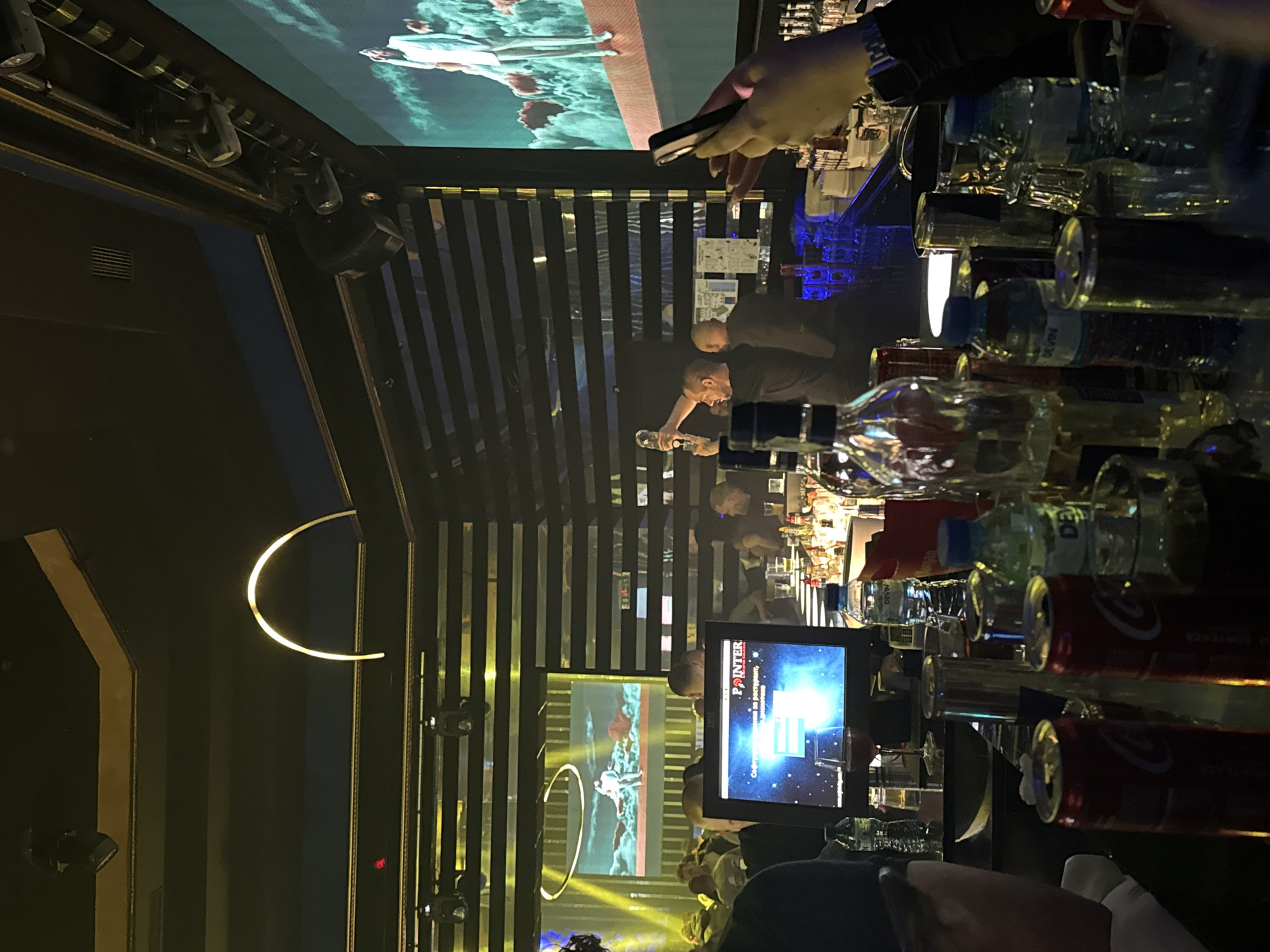 The image depicts the interior of what appears to be a nightclub or bar in Goce Delcev, Bulgaria, at night. A bar is in the foreground filled with bottles and cans. Bartenders are working behind the bar, serving customers. Several television screens are visible displaying different images. There are other people in the background, presumably patrons of the establishment. The environment is dark, with lighting creating ambiance.