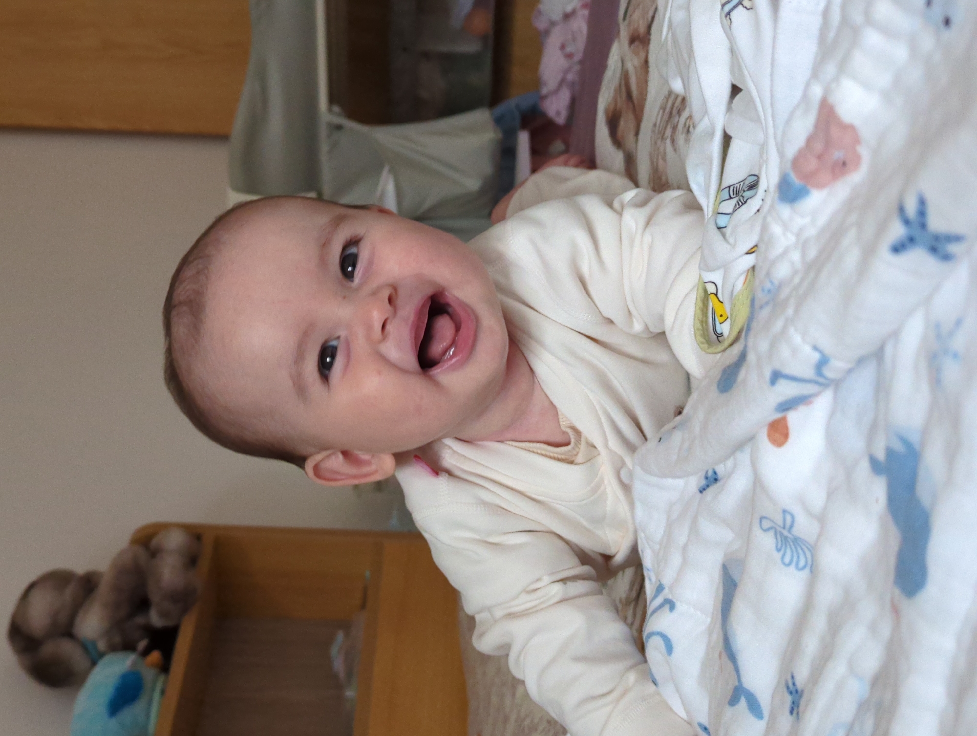 A baby is photographed indoors. The baby is light-skinned and is lying on a white blanket with a pattern of blue sea creatures, wearing a cream-colored, long-sleeved shirt. The baby is looking at the camera and has their mouth open, showing their tongue. A wooden structure is visible in the bottom left corner, containing stuffed animals. A metal bassinet is behind the baby, and a tan-colored wooden wall is visible in the upper left. There is no information to indicate this photo was taken in Shunde District, China.