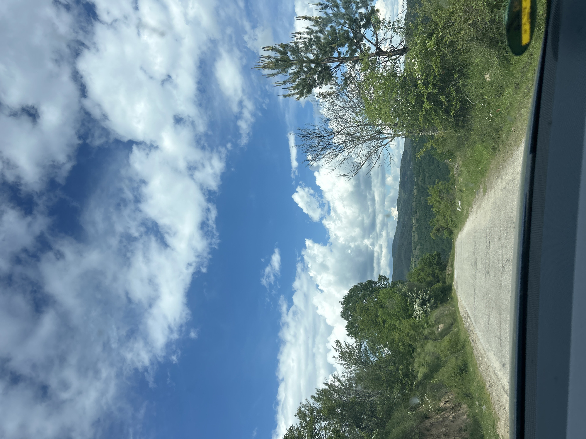 Captured through a car window, the image presents a view of a narrow, asphalt road winding through a verdant, hilly landscape. The sky, a vibrant blue, is adorned with cumulus clouds. Green vegetation lines the roadside, with trees scattered throughout. A mountain range appears faintly in the distance. The scene depicts the Dolen area in Bulgaria.