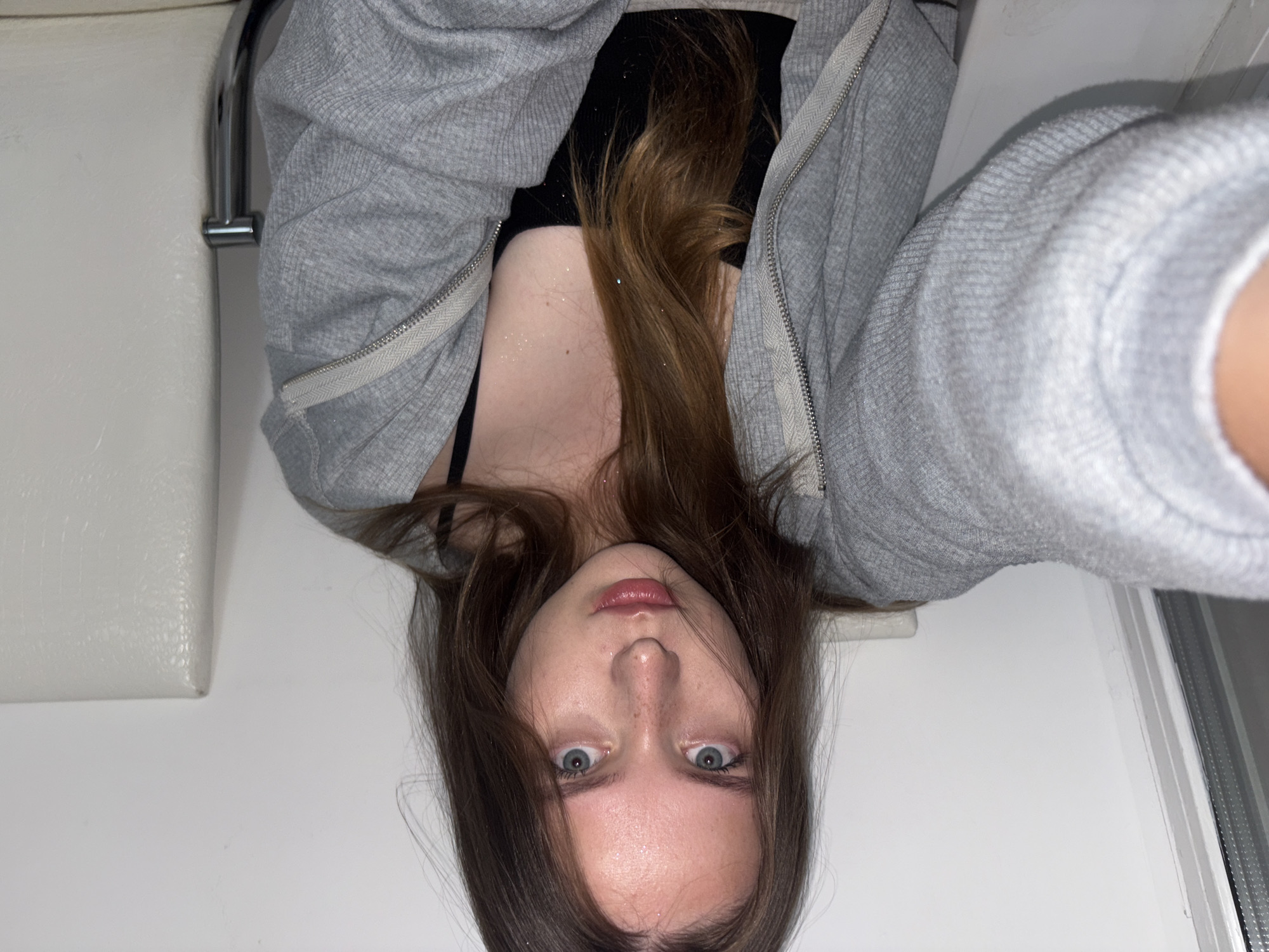 A young Caucasian woman with medium-length brown hair and blue eyes is seated, holding a camera to take a selfie. She is wearing a grey zippered hoodie over a black tank top. Her face is looking directly at the camera, with a neutral expression. She has freckles across her nose and cheeks. The background is a plain white wall, with a white chair visible to her right. The lighting is even and suggests an indoor setting. The image appears to be taken in a residential or public indoor space, possibly a waiting area or a room within a building. There are no specific visual cues that would definitively place this image in Chaikovsky, Russia, or any other city.