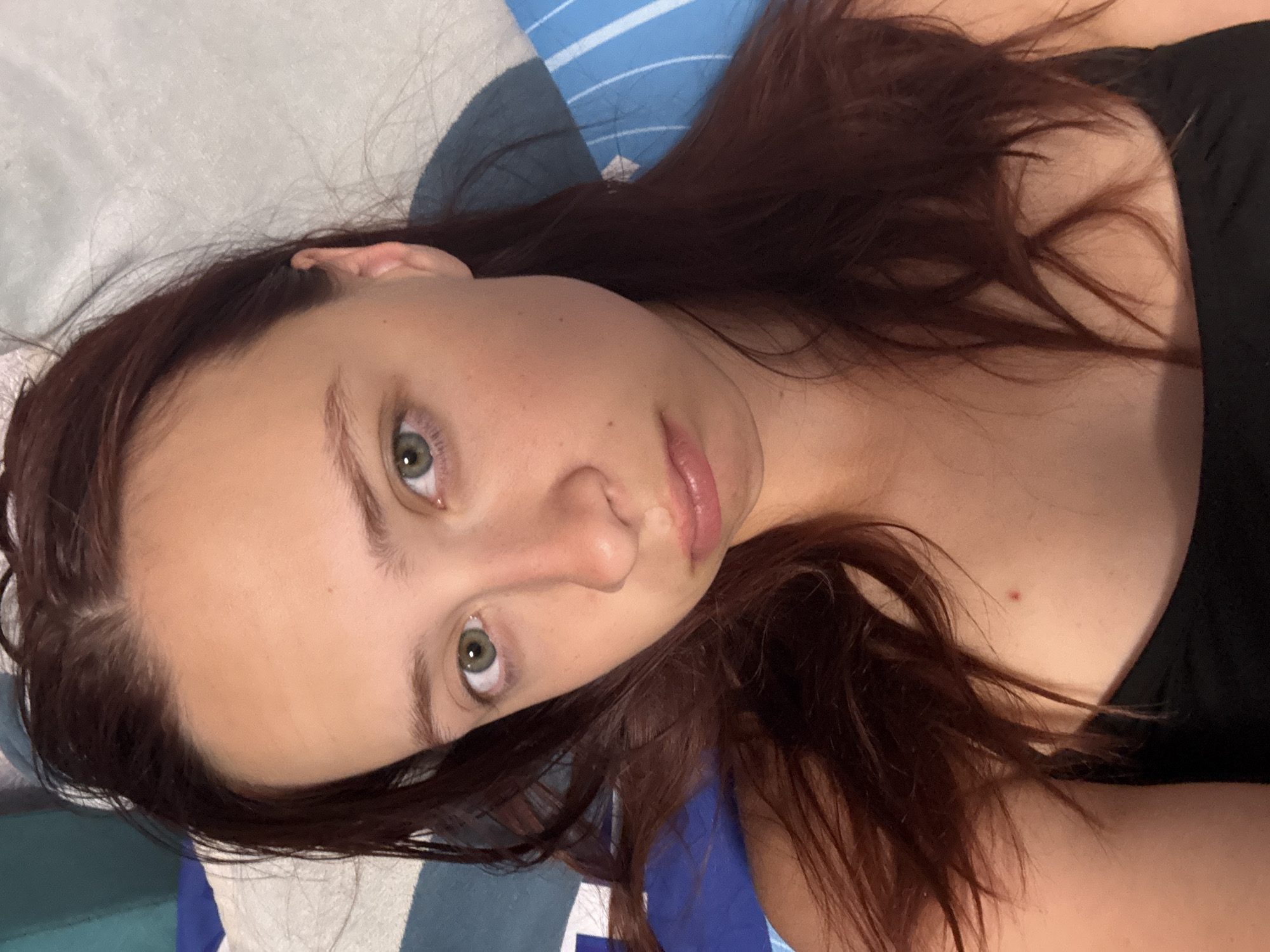 A fair-skinned woman with auburn hair and green eyes is lying on her back on a blue and white striped pillow. She is wearing a black top. The location, Прикамский, Russia, cannot be determined from this image.