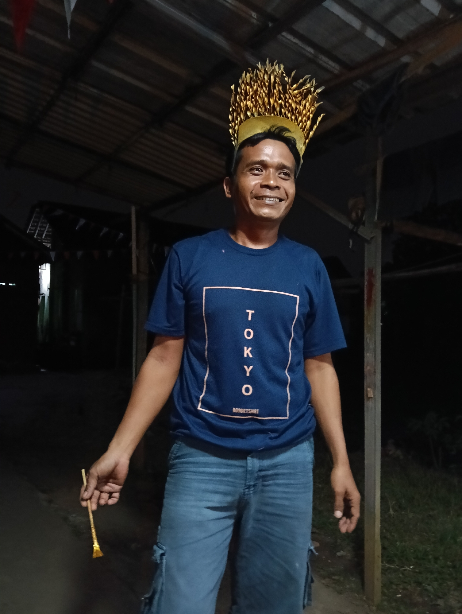 A man stands outdoors at night, possibly in a backyard or an unpaved area, wearing a blue t-shirt with "TOKYO" printed vertically inside a square outline and blue jeans. He has a gold, leaf-shaped headpiece or crown on his head. In his right hand, he holds a small, golden object that resembles a wand or a miniature broom. The background shows a dark, open structure with a corrugated roof and some greenery. There are faint triangular flags strung across the structure. The lighting suggests it is evening or night. The location is a city in Tasikmalaya, Indonesia.