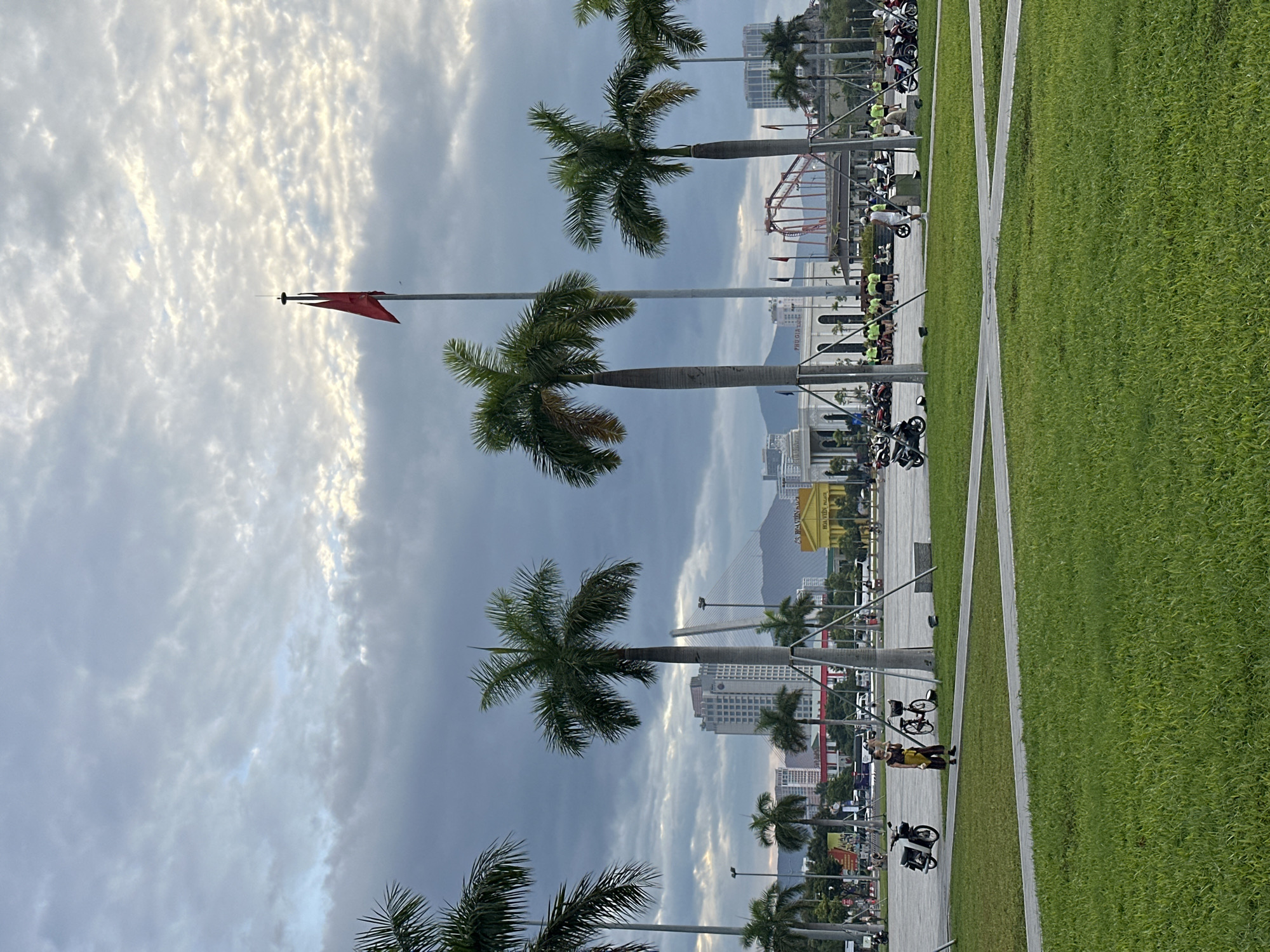 The image depicts a wide, open plaza in Da Nang, Vietnam, likely near a waterfront given the presence of palm trees and a hazy sky suggesting a humid climate. In the foreground, a meticulously maintained green lawn is intersected by light-colored pathways. A single individual, possibly a young person, stands on one of these paths, holding a small object and looking towards the right side of the frame. To their left, a motorcycle is parked. Further in the background, several other motorcycles are visible, some in groups, indicating a public gathering or transportation hub.
The middle ground is dominated by tall palm trees, characteristic of tropical urban landscapes. Beyond the palm trees, a collection of buildings rises, including a prominent yellow structure with signage that reads "HOA VIEN PALACE." Other multi-story buildings and a suspension bridge can be seen in the distance, hinting at the city's modern infrastructure. A tall flagpole with a red flag is positioned near the center of the image, against a cloudy sky. The clouds are varied, with patches of lighter sky breaking through the darker, more ominous ones, suggesting either early morning or late afternoon light. The overall impression is one of a bustling urban environment in a tropical setting, with a sense of public space and activity.