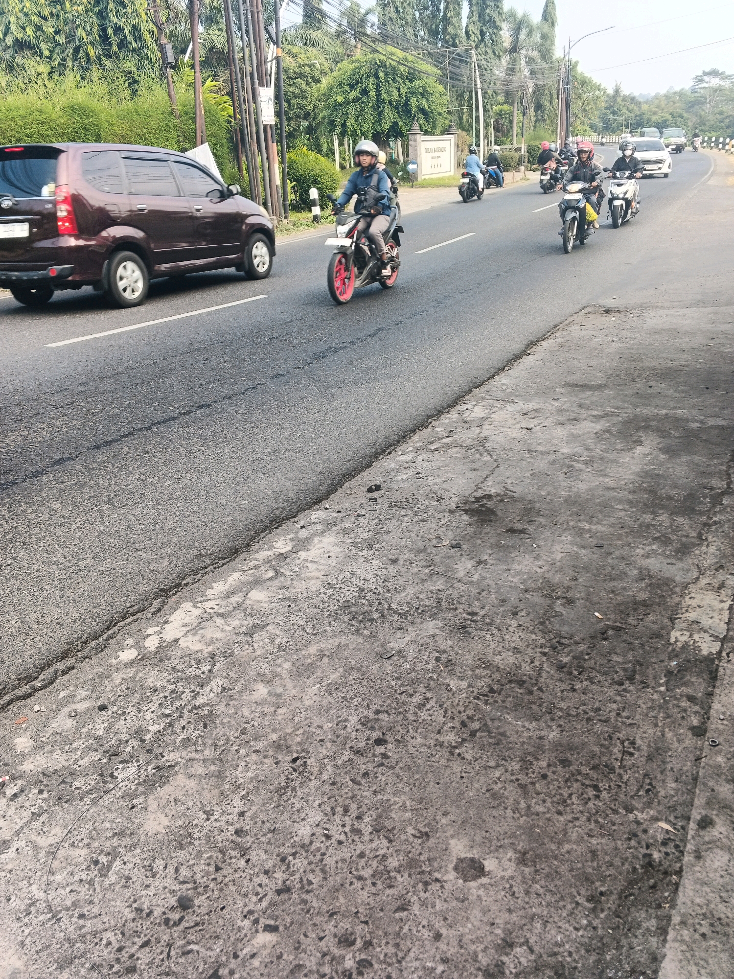A wide shot captures a busy street in Ungaran, Indonesia, likely on Jalan Diponegoro. A dark brown Avanza minivan is seen in the left lane, with its rear facing the camera. In the right lane, several motorcycles are traveling in the same direction. The first motorcycle, closer to the foreground, is a red and black model ridden by a person wearing a blue jacket and a white helmet. Further ahead, a white motorcycle with a rider in a black jacket and a dark helmet is visible. Beyond them, a yellow motorcycle is also in motion, with its rider wearing a red helmet. The road is asphalt, with white lane markings. On the right side of the road, a concrete sidewalk is visible, showing signs of wear and tear. The background is filled with lush green trees and foliage, with utility poles lining the side of the road. The sky is bright, suggesting a daytime scene. A sign with Indonesian text is visible on the left side of the road, indicating a business or institution.