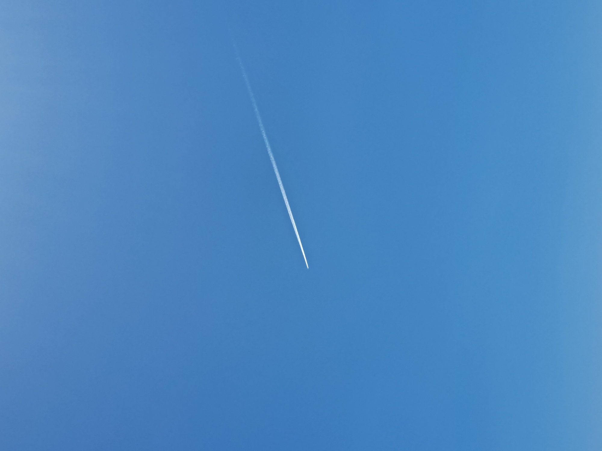 A contrail streaks diagonally across a clear blue sky. The contrail is white and thin, originating from an unseen aircraft. The sky is uniformly blue with slight variations in tone. The scene is outdoors. The location is unknown.