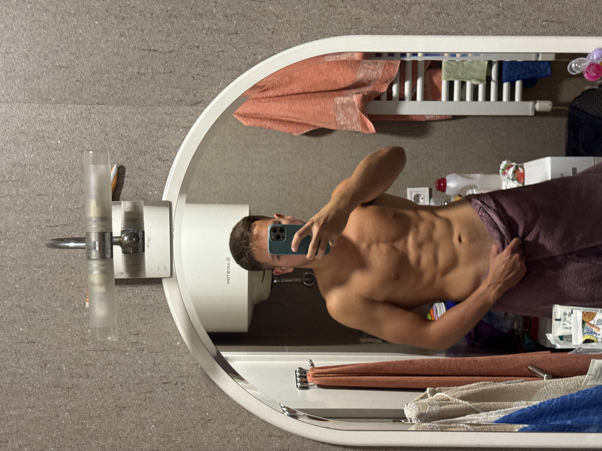 A young man with well-defined abdominal muscles and tanned skin, wearing a purple towel wrapped around his waist, is taking a mirror selfie in a bathroom. He holds a teal smartphone in his right hand. The bathroom has grey tiled walls and a large arched mirror. A white water heater is visible behind him in the reflection. To the left of the mirror, several towels of different colors are hanging on hooks. To the right, a white heated towel rack holds folded towels. The scene suggests a private residential bathroom, possibly in a hotel or apartment in Budva, Montenegro.