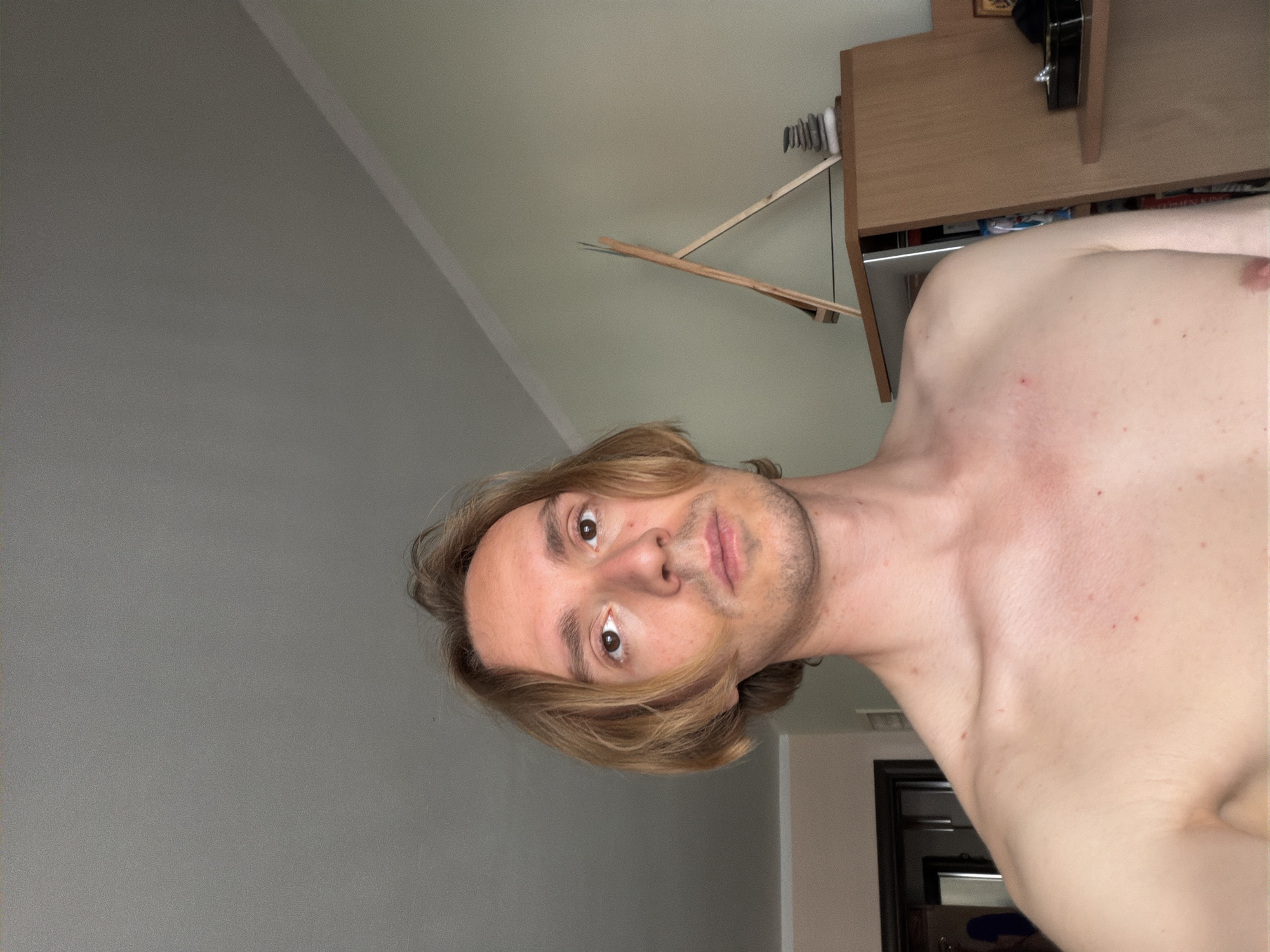 The image shows a medium shot of a shirtless young man with light brown hair, standing indoors. He is looking directly at the camera with a neutral expression. The background includes a wall, a corner of a room, and furniture. There are no explicit indicators of the city of Budva, Montenegro, within the image itself. The man's skin has some small red spots. A wooden easel is visible behind him, and a stack of stones rests on a shelf.