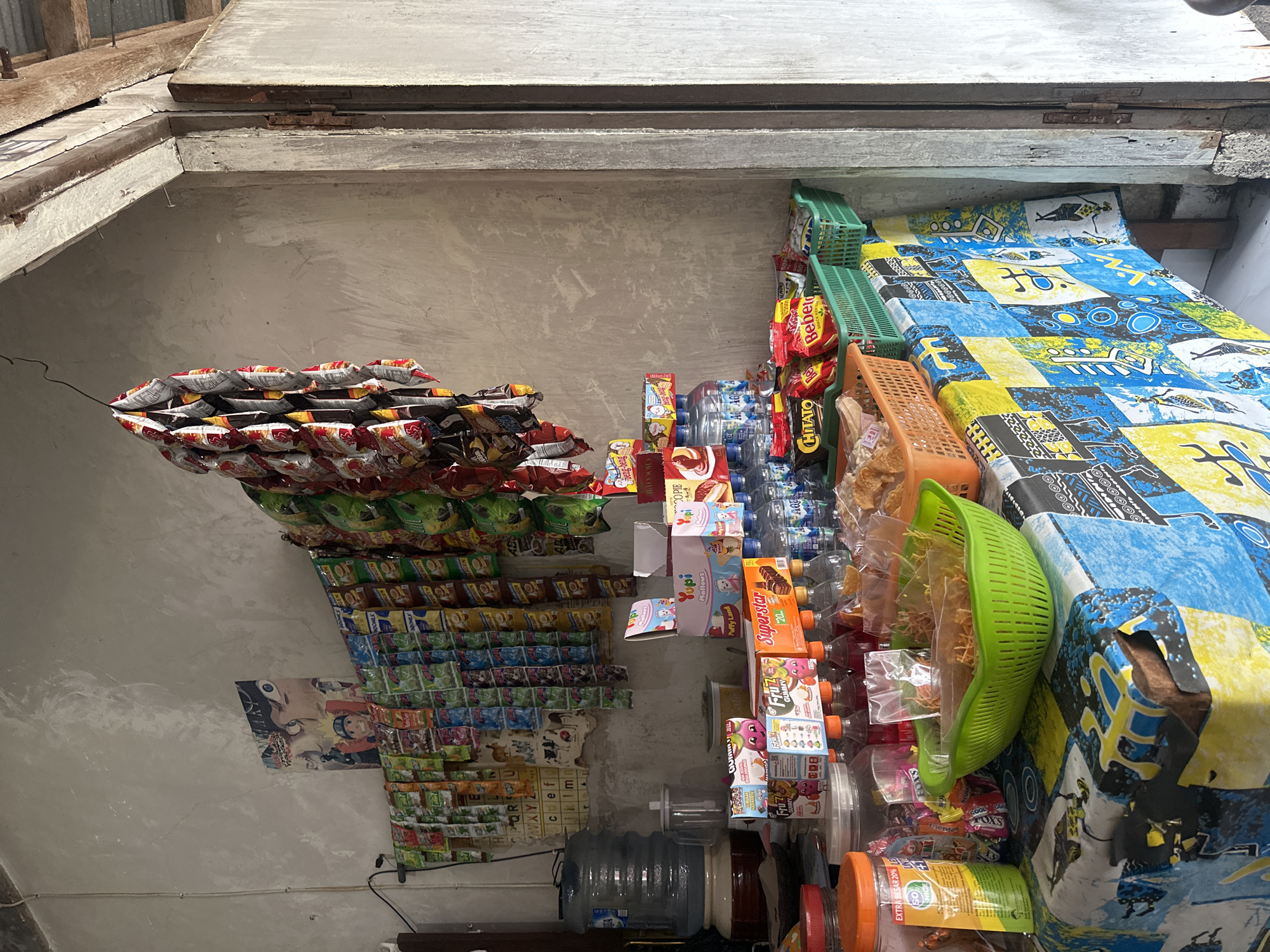 A street-side kiosk in an urban environment displays a variety of snacks and beverages for sale. The kiosk features a variety of products including bottled water, sodas, chips, and candies. Some items are hung on display racks, while others are placed on shelves and a table covered with a brightly colored patterned cloth. A large water dispenser is visible on the left side of the kiosk. The background consists of a concrete wall and a wooden doorway. The overall scene depicts a small, informal retail setup.