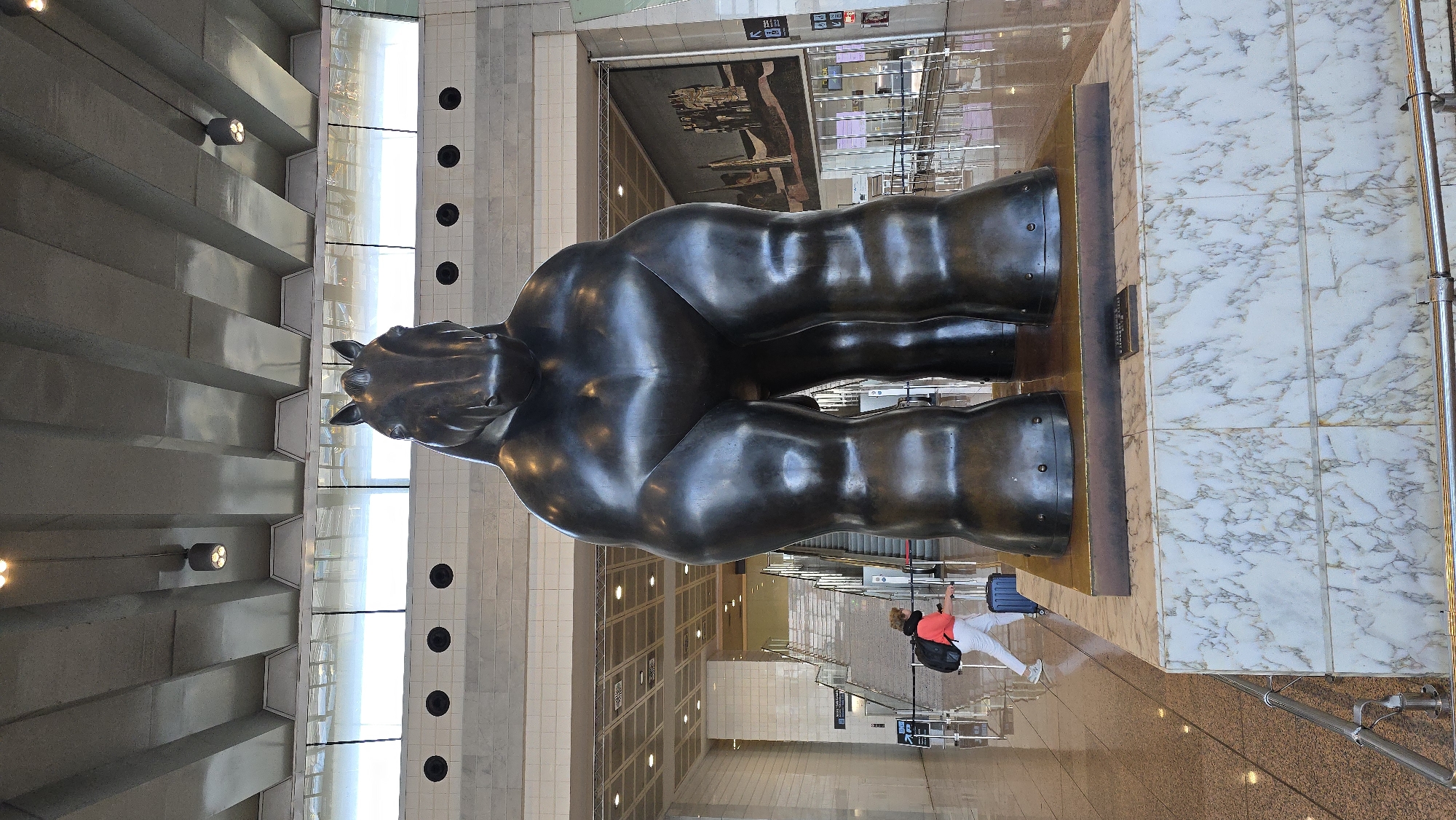 A large, dark bronze sculpture of a horse, characterized by exaggerated muscular forms, stands prominently in the foreground of a modern indoor public space. The sculpture is mounted on a tiered base, the lower portion of which is clad in white marble with grey veining. The background reveals an airport terminal environment. To the left, a woman wearing a coral jacket, white pants, and sneakers is walking away from the viewer, pulling a dark blue rolling suitcase and carrying a black backpack. She is positioned near a set of escalators and stairs. In the distance, a large mural or artwork is visible on the wall, depicting several figures. The space features high ceilings with exposed structural elements and lighting fixtures, and a reflective floor. The overall impression is that of a contemporary art installation within a busy transportation hub. This sculpture is located in Terminal 1 of Barcelona-El Prat Airport in El Prat de Llobregat, Spain.